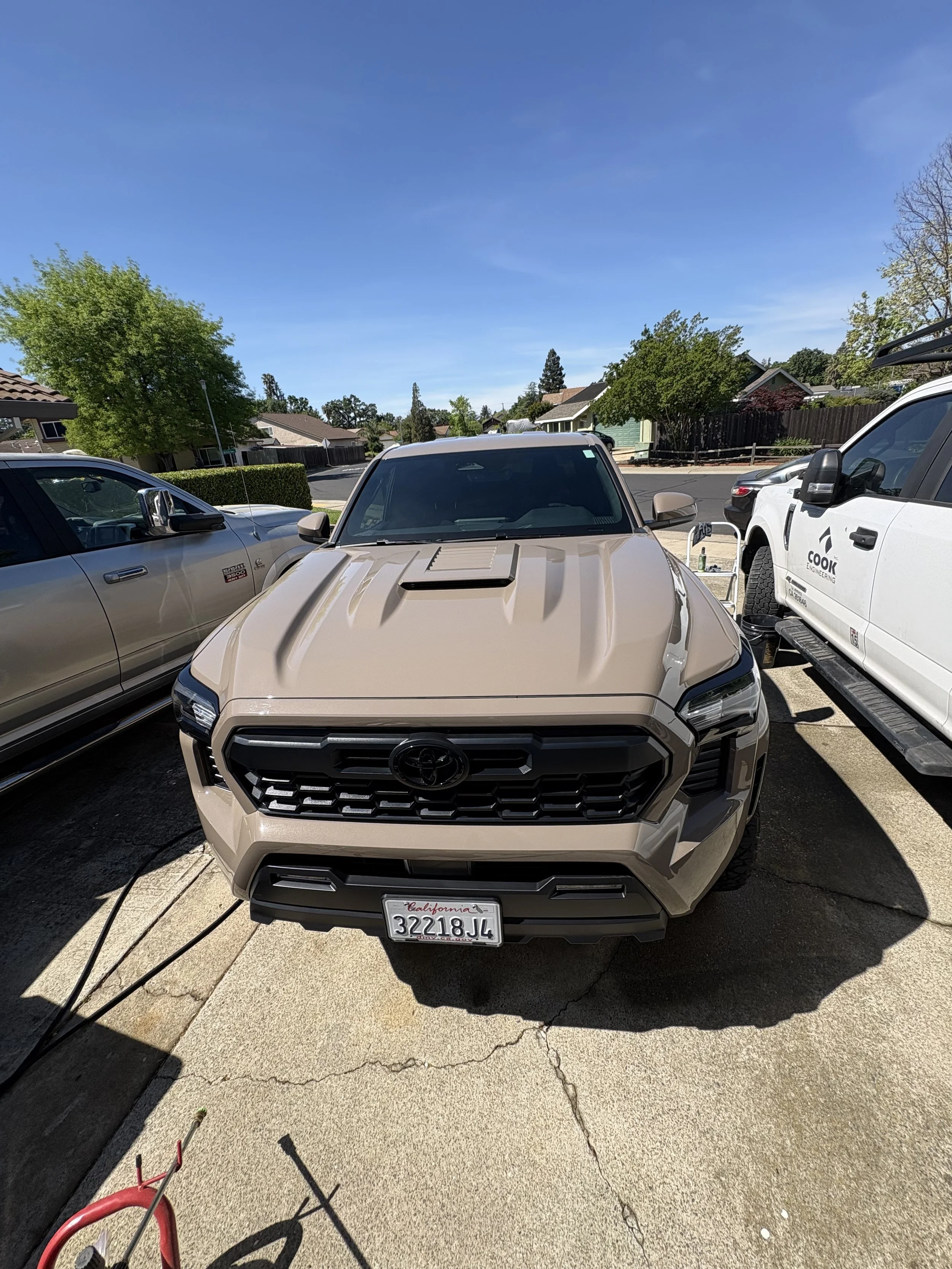 Front view of a beige Toyota pickup truck with California license plates parked on a driveway, with other vehicles and houses in the background, under a clear blue sky.