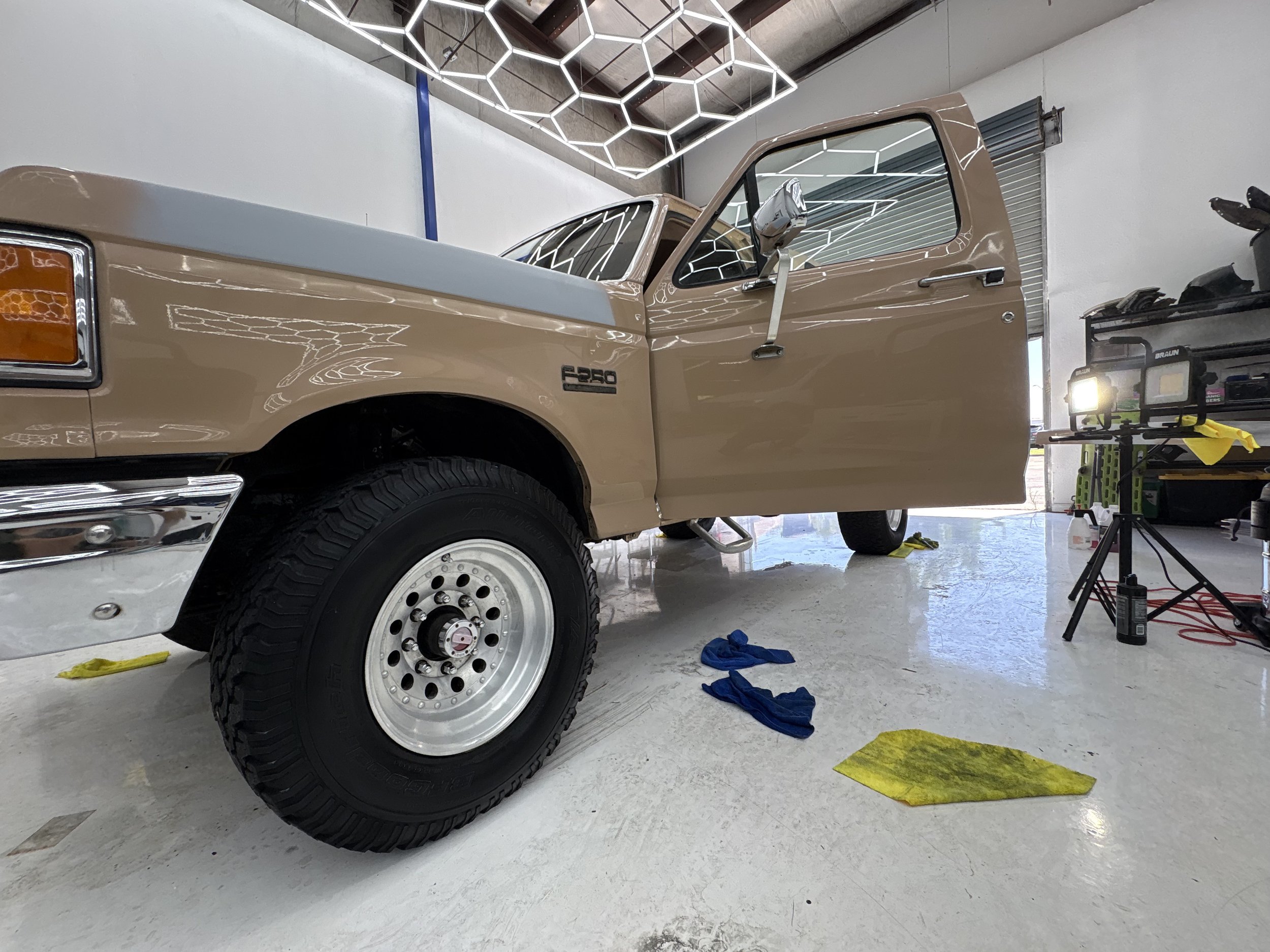 A beige pickup truck inside a garage undergoing repairs or restoration, with tools and equipment nearby, under a geometric lighting installation on the ceiling.