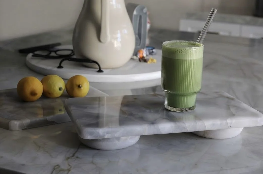 A marble tray holding a green smoothie with a straw, three yellow lemons on a small marble cutting board, on a marble countertop in a kitchen with a teapot and other items in the background.