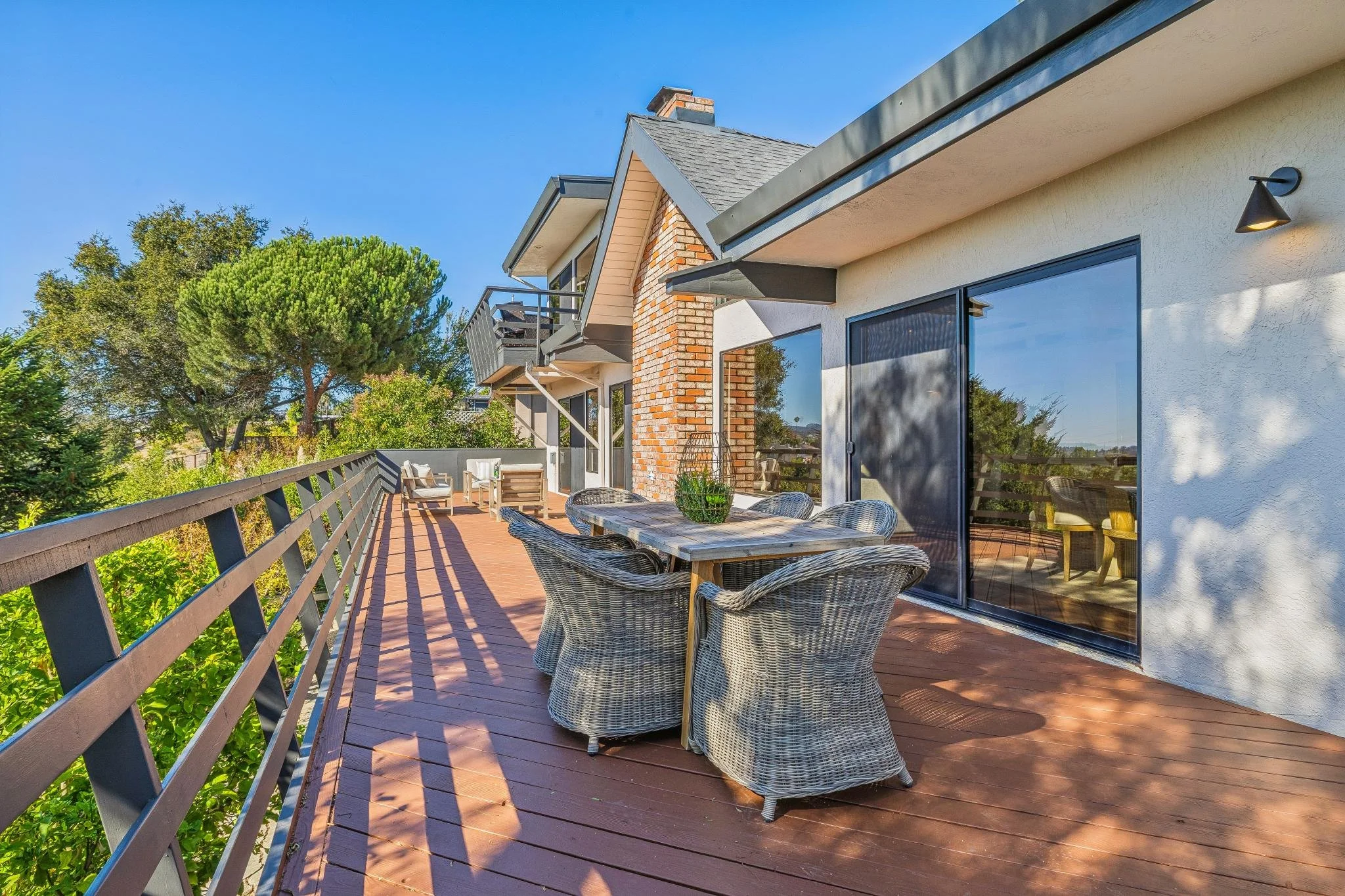 Outdoor balcony with wicker furniture, wooden dining table, and chairs, surrounded by trees and residential buildings under a clear blue sky.