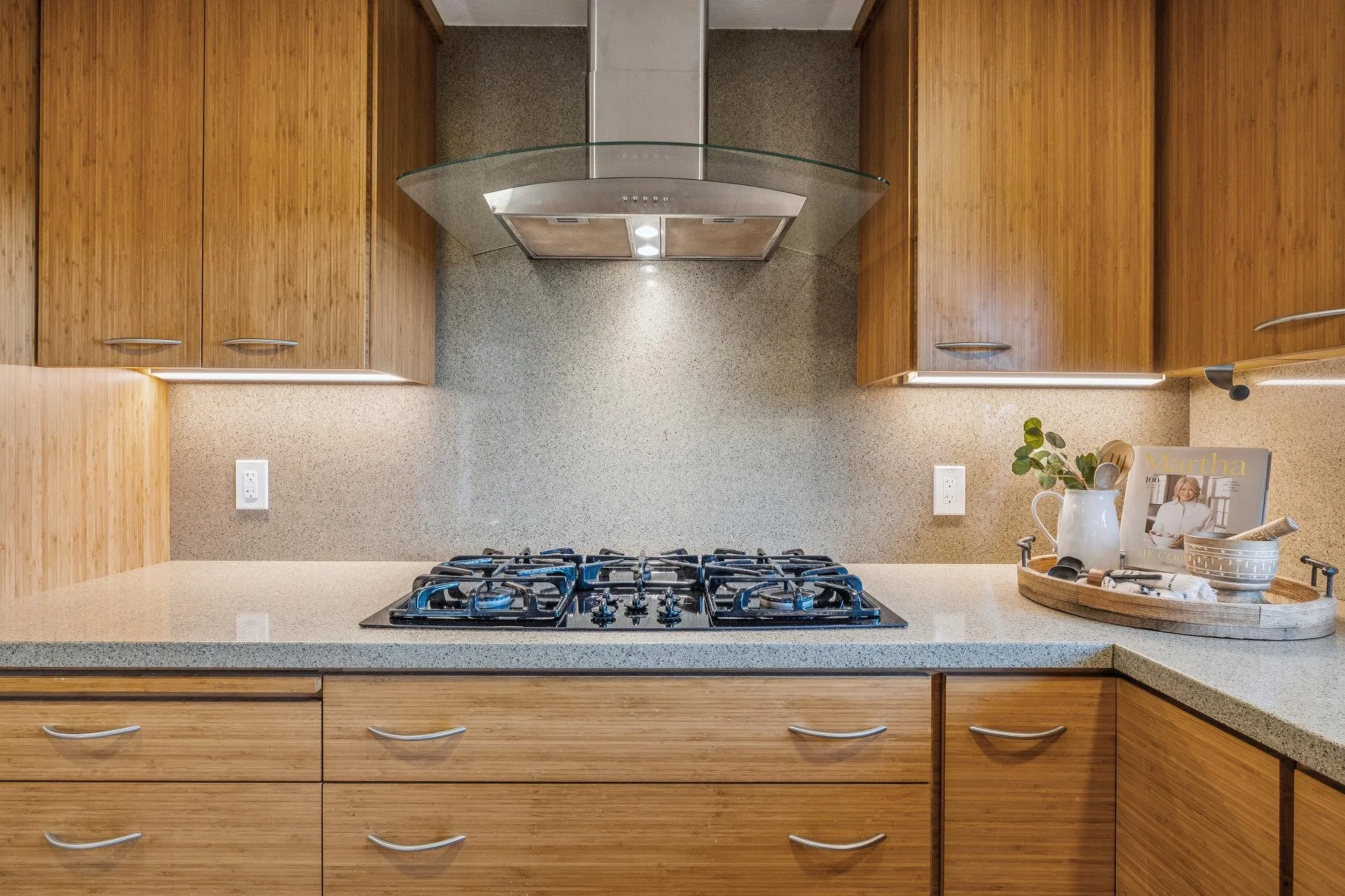 A kitchen countertop with a built-in gas stove, wooden cabinets above and below, and a beige speckled backsplash. On the right side, there is a white mug, a magazine featuring Martha Stewart, a small plant, and a round tray with various items, set on