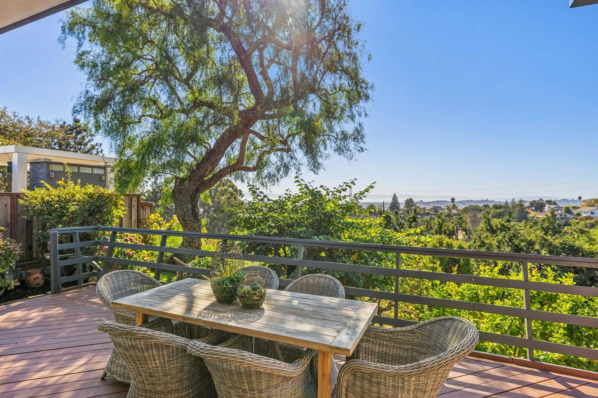 A backyard patio with a wooden table and woven chairs, lush green trees, and a view of the city in the distance under clear blue sky.