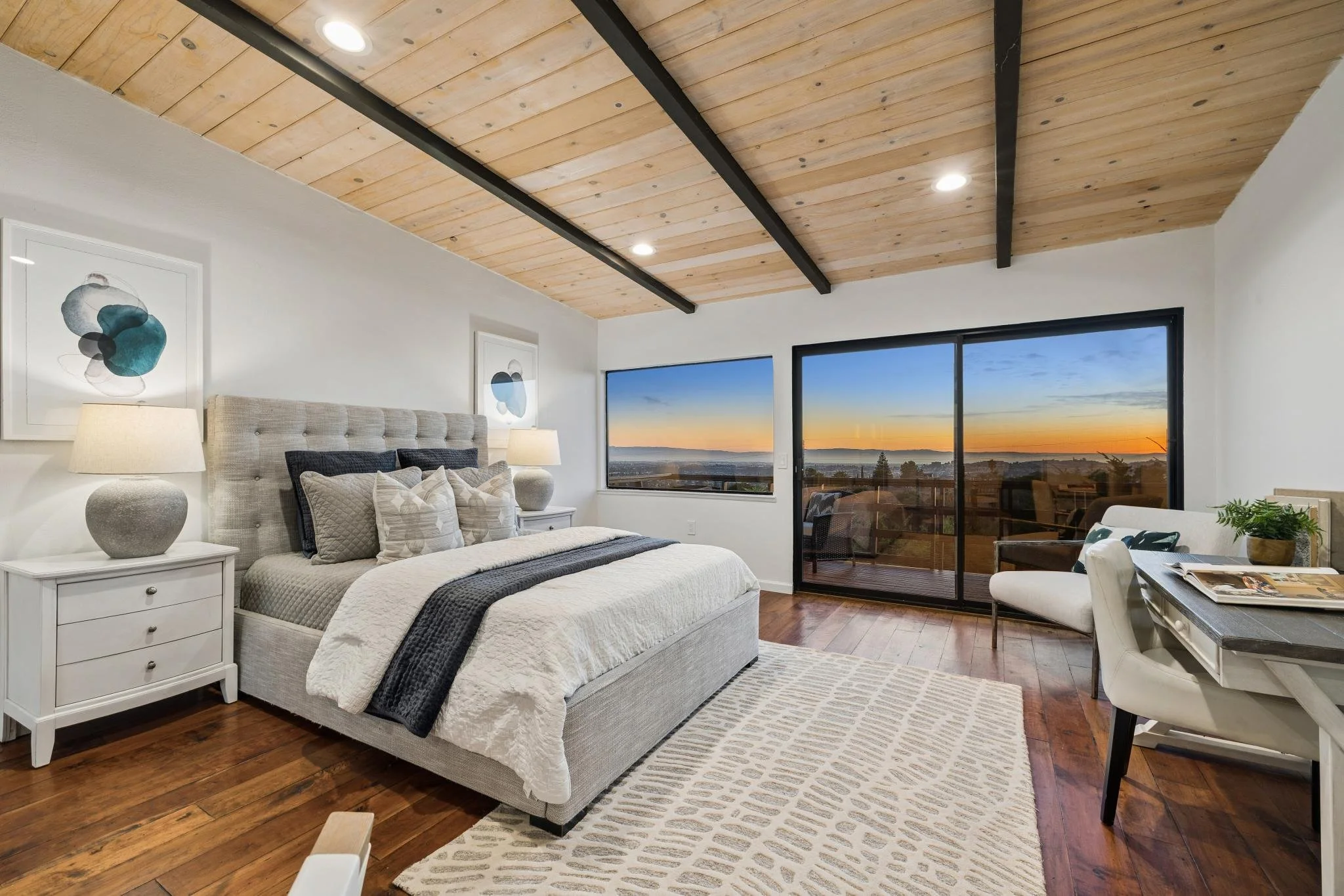 Modern bedroom with wooden ceiling, large window showing a sunset view, white bed with gray and black pillows, two matching bedside lamps, abstract artwork on the wall, and a sitting area with a table and chair.