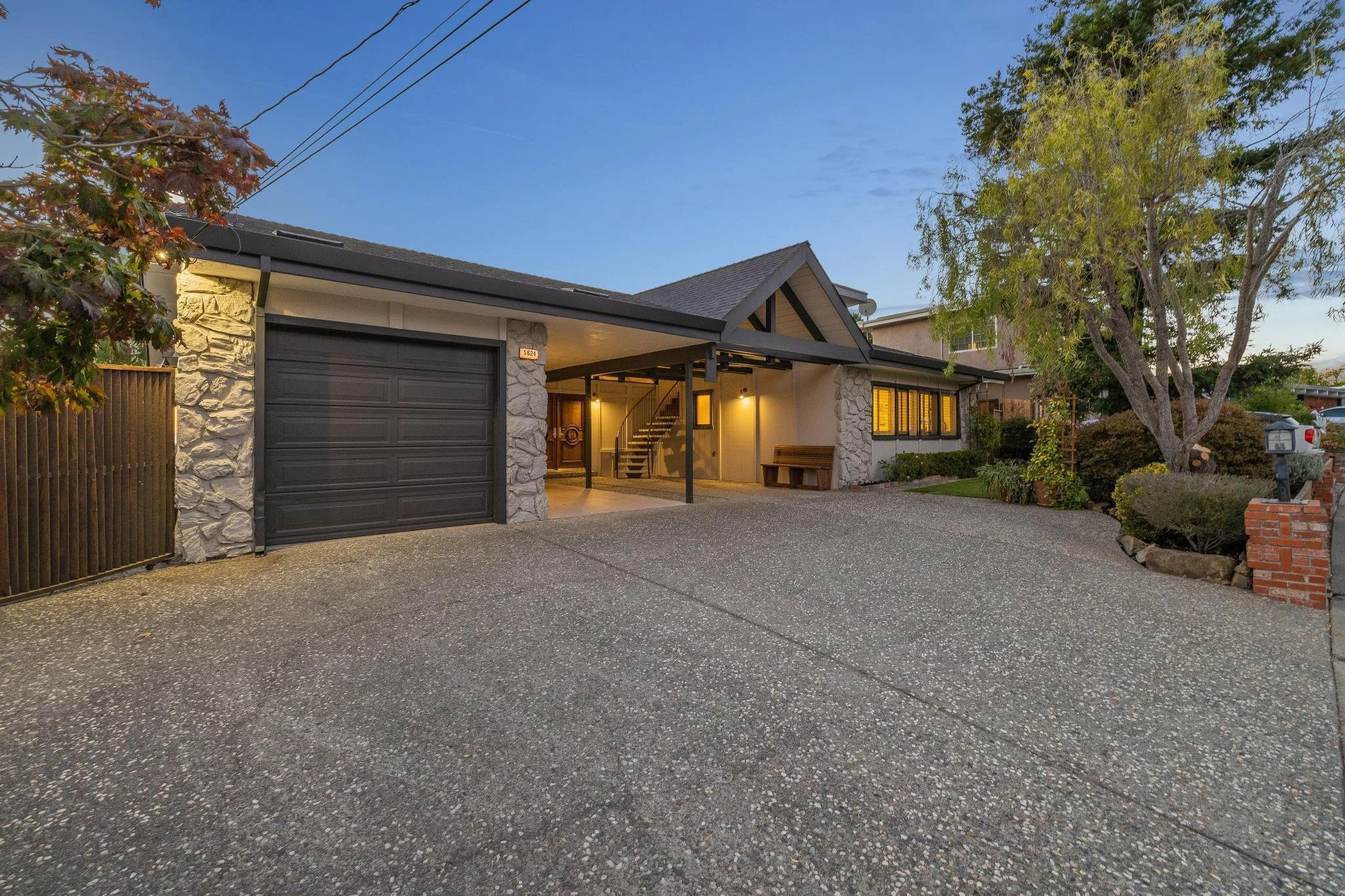 Front view of a modern house during twilight with a stone and siding exterior, large windows with yellow lighting, attached garage, and a short driveway surrounded by a landscaped yard with trees and shrubs.