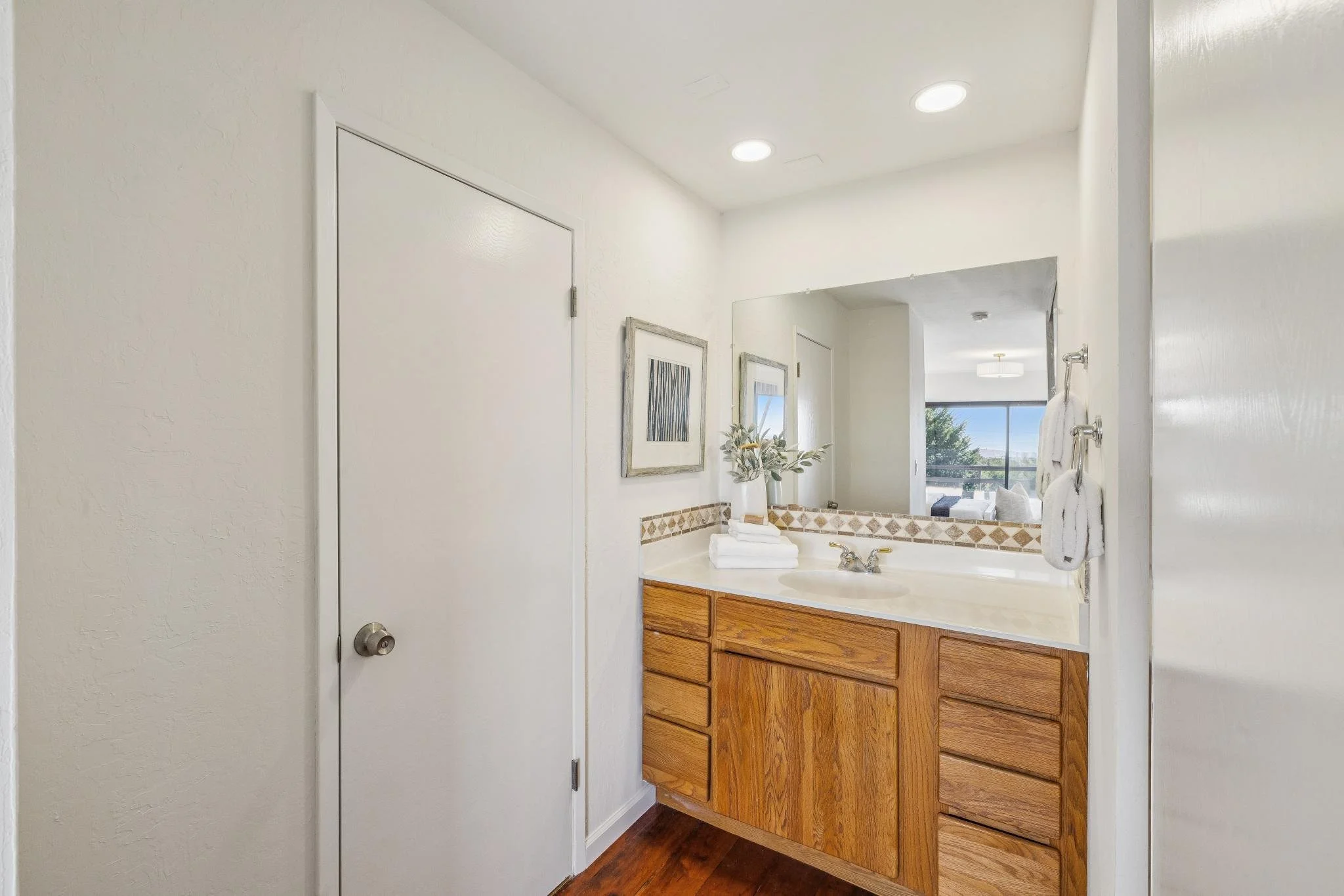 Bathroom with a wooden vanity, a mirror, framed artwork, a plant, and towels, with a window showing a view outside.