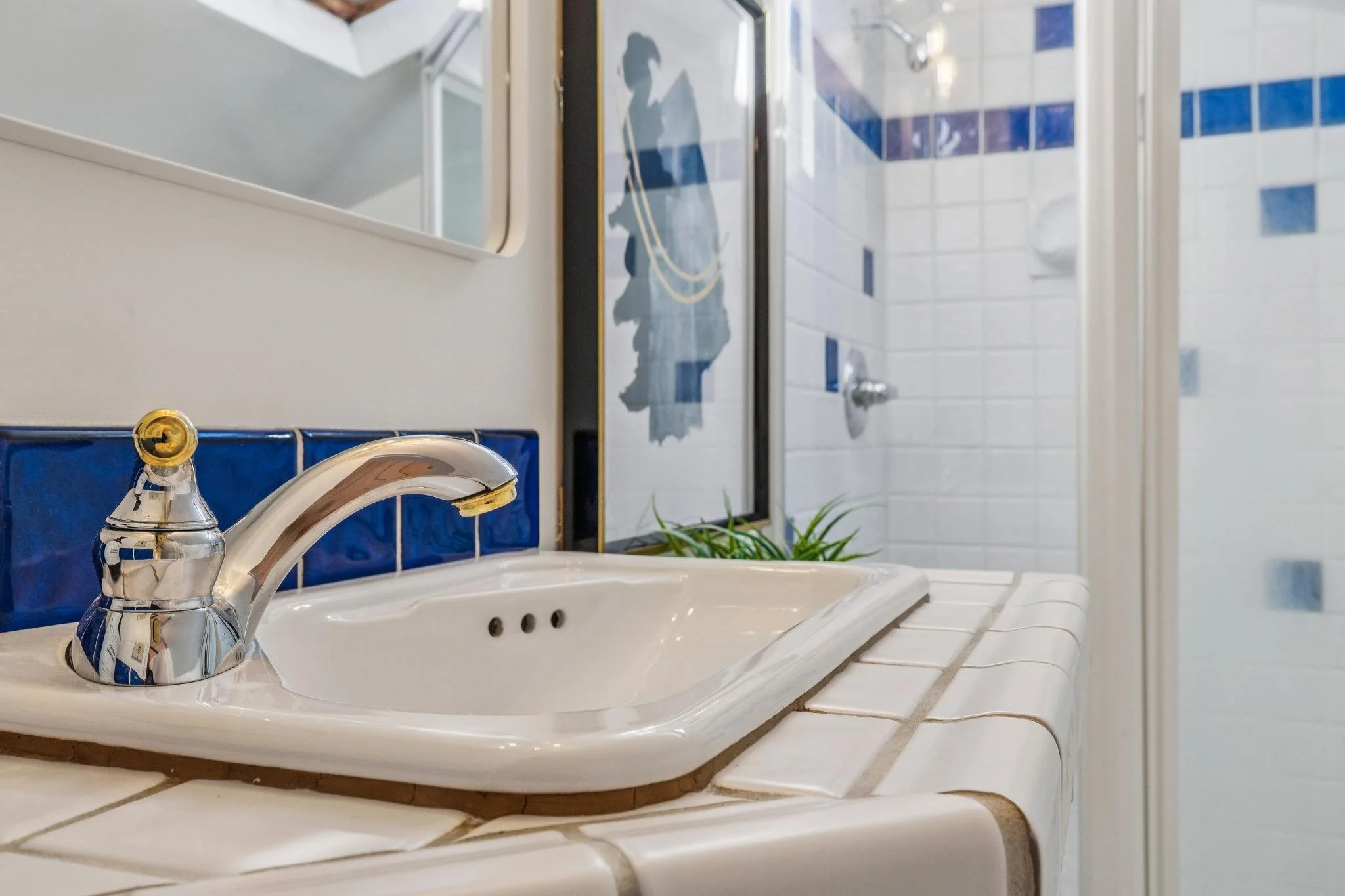 Bathroom sink with chrome faucet on a white tiled countertop, mirror and shower area with white tiles and blue accents in the background.