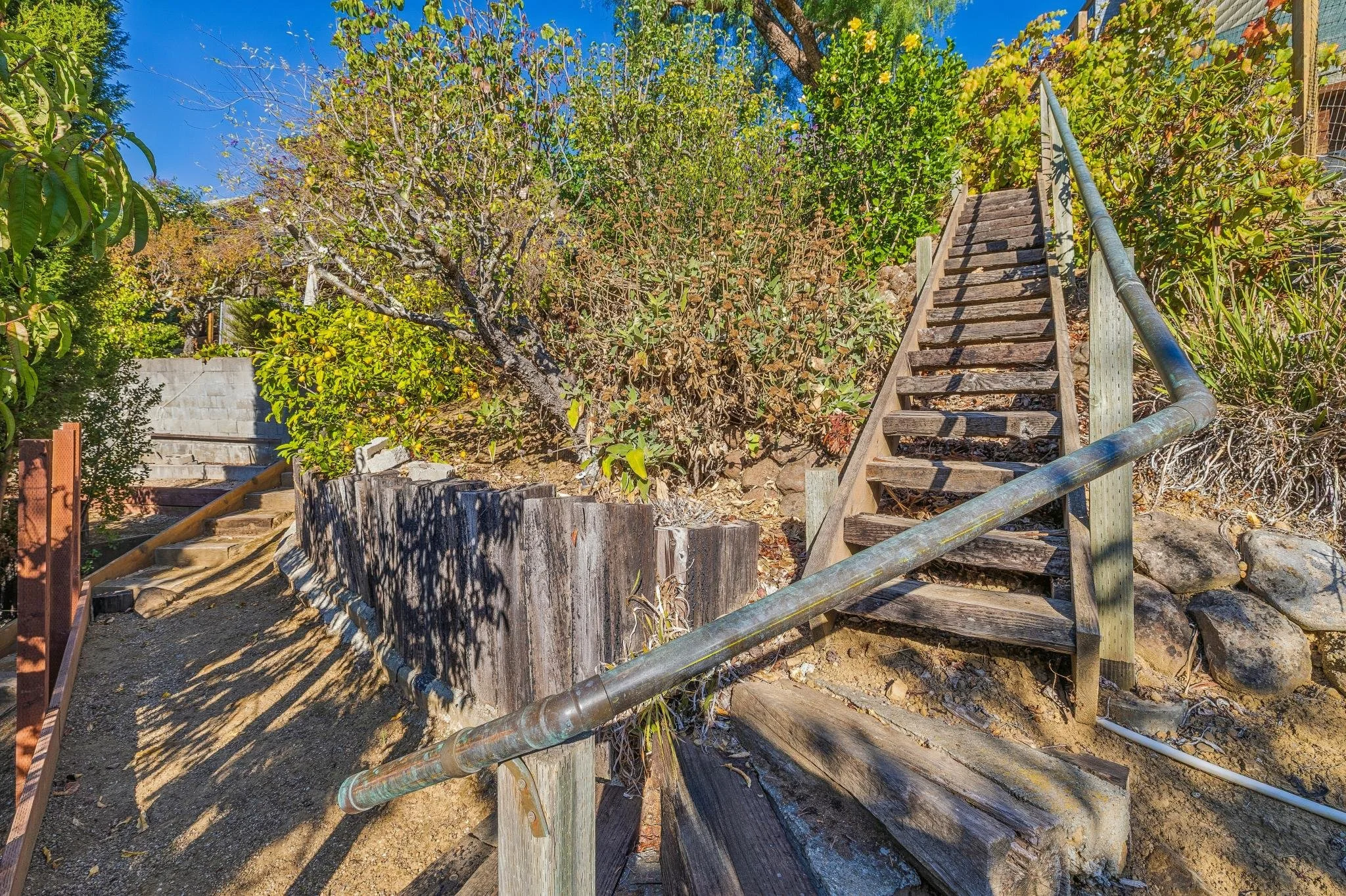 Wooden stairs with metal handrails leading up a hill surrounded by green bushes and trees, with a clear blue sky overhead.