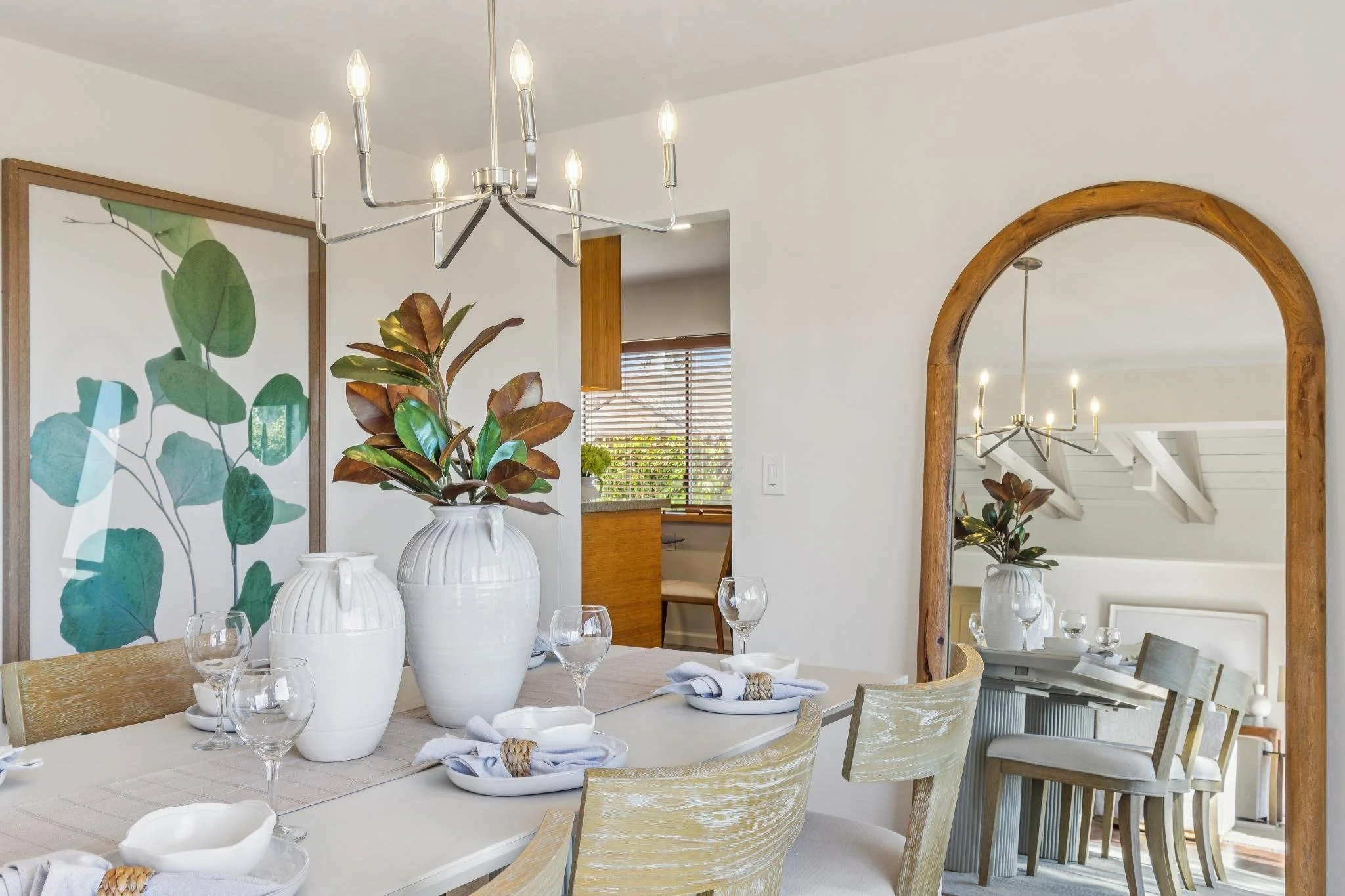 A dining room with a white table set with glasses and white bowls, decorated with large white vases containing green and brown leaves, a large mirror with a wooden frame, and a modern chandelier with multiple lights hanging from the ceiling.