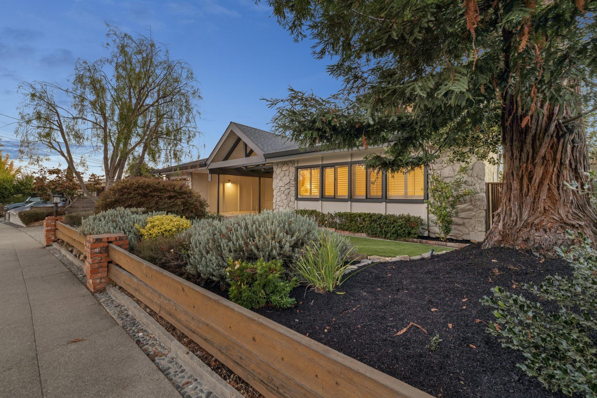 A house with large windows and a stone exterior, surrounded by trees and bushes, under a blue sky during dusk.