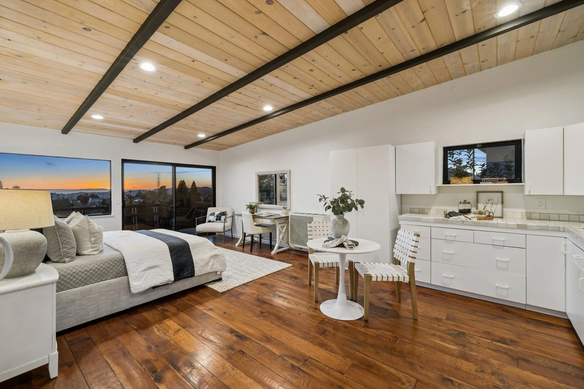 Living room with a wooden ceiling, large windows showing sunset, white walls, beige sofa with pillows, small white table, and kitchen area with white cabinets and countertops.