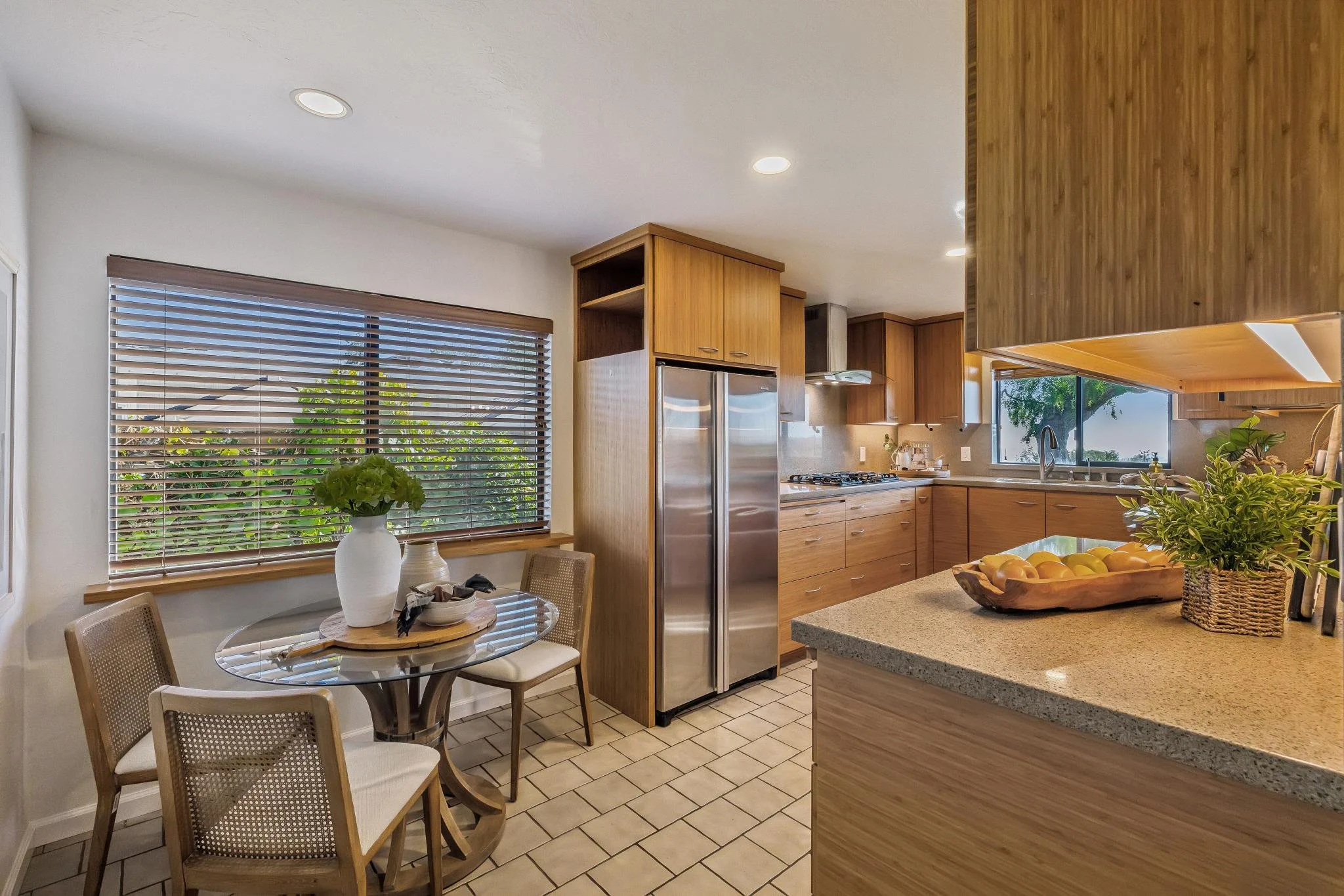 View of a kitchen with a round dining table and four chairs, a large window with blinds showing green foliage outside, wooden cabinets, a stainless steel refrigerator, a kitchen island with a fruit bowl and plants, and a ceiling with recessed lightin