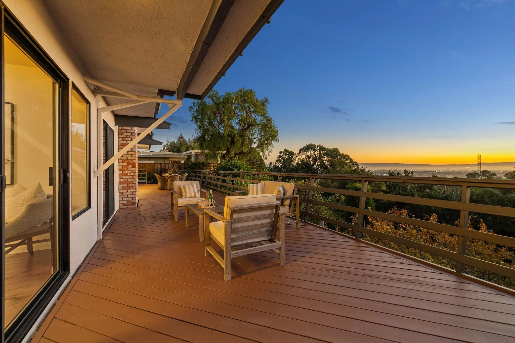 Balcony with outdoor seating, view of trees, sunset sky, and distant landscape.