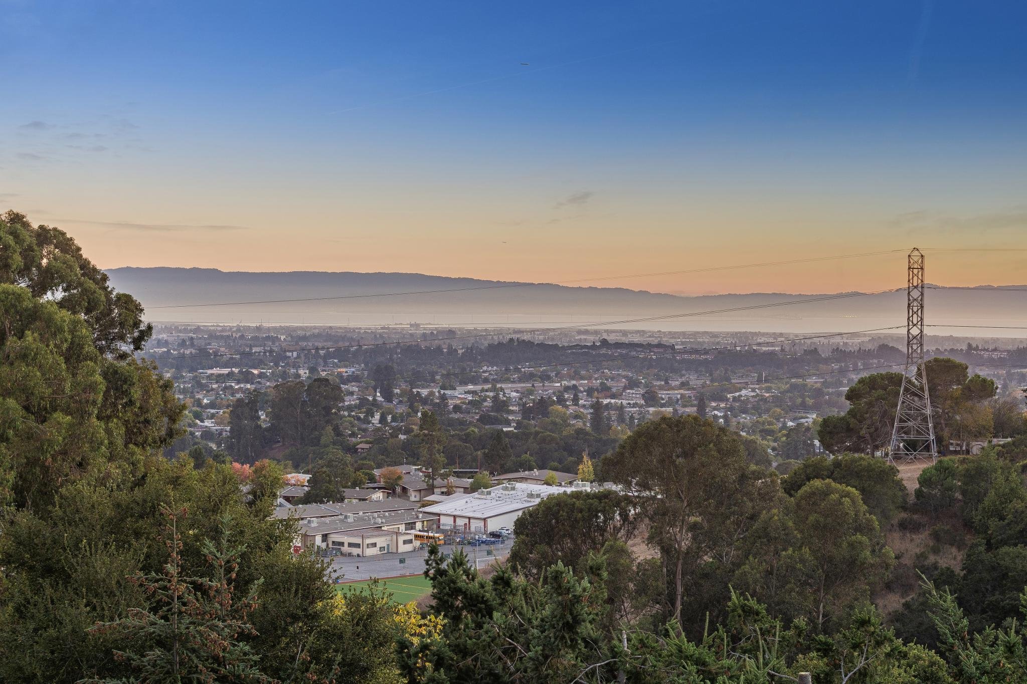 A scenic view of a city and suburban area with trees in the foreground, industrial buildings, a large power line tower, and mountains in the background under a clear sky during sunset or sunrise.