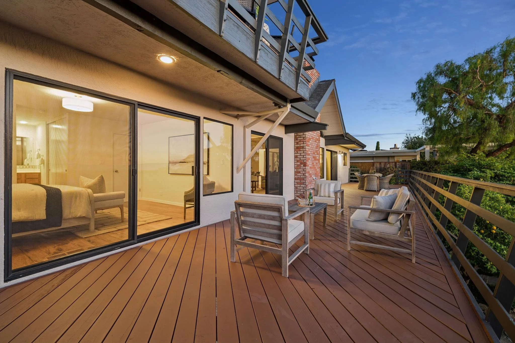 A spacious outdoor balcony with wooden decking, white outdoor furniture including chairs and a table, large glass sliding doors revealing a living room interior, and a green leafy tree on the right side, under a blue evening sky.