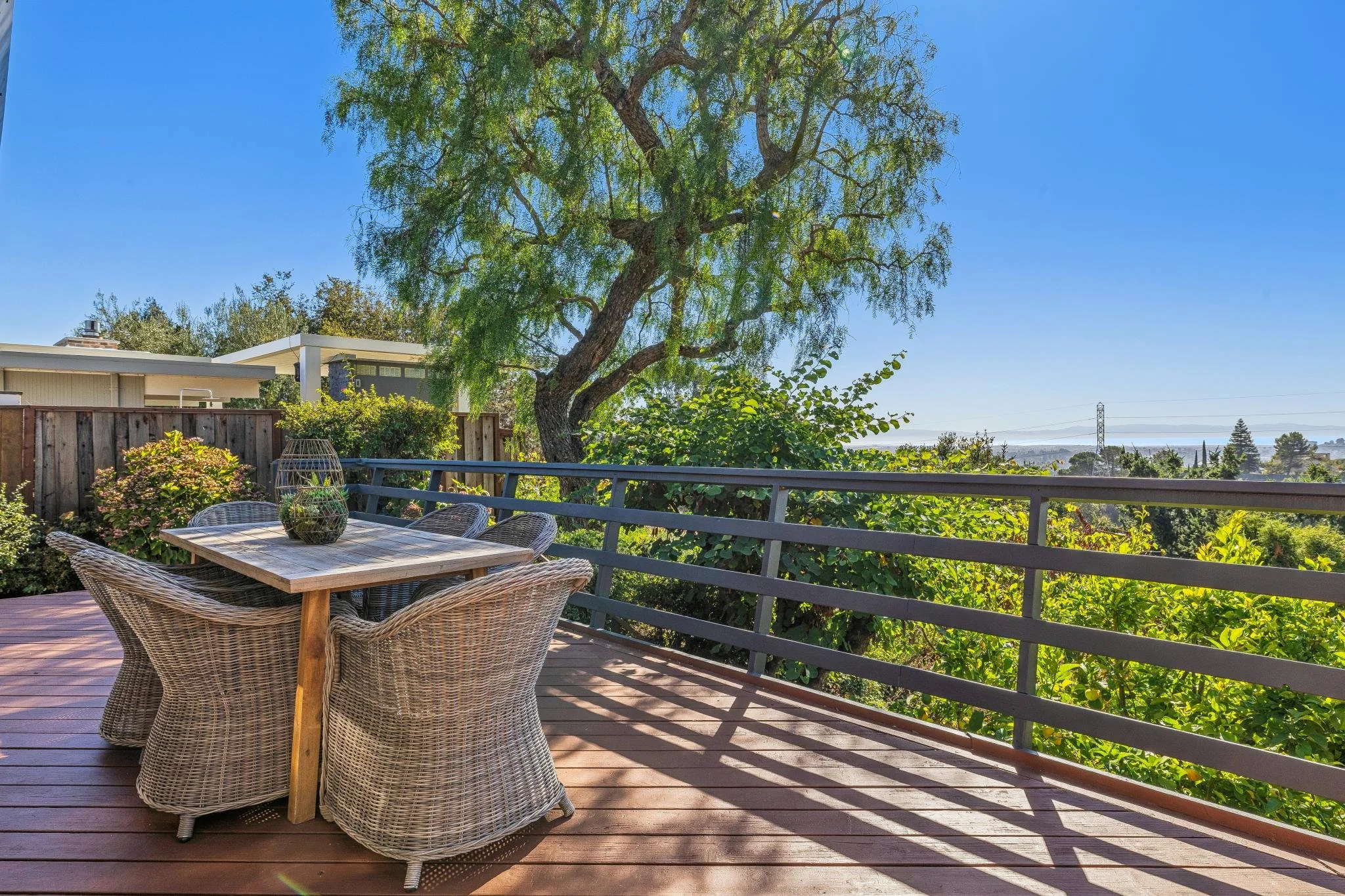 Outdoor wooden deck with wicker chairs and a table, overlooking a lush green landscape with large trees, a fence, and distant houses under a clear blue sky.