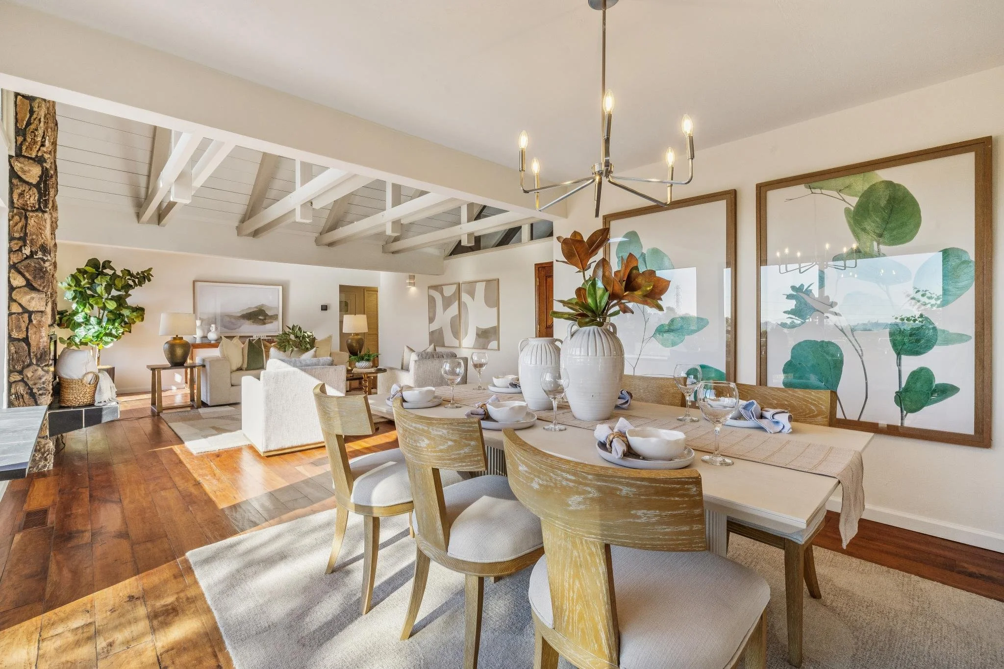 Interior of a modern dining and living area with a wooden dining table, white vases with large green plants, a chandelier, large framed artwork of leaves, and a sitting area with sofas and lamps.
