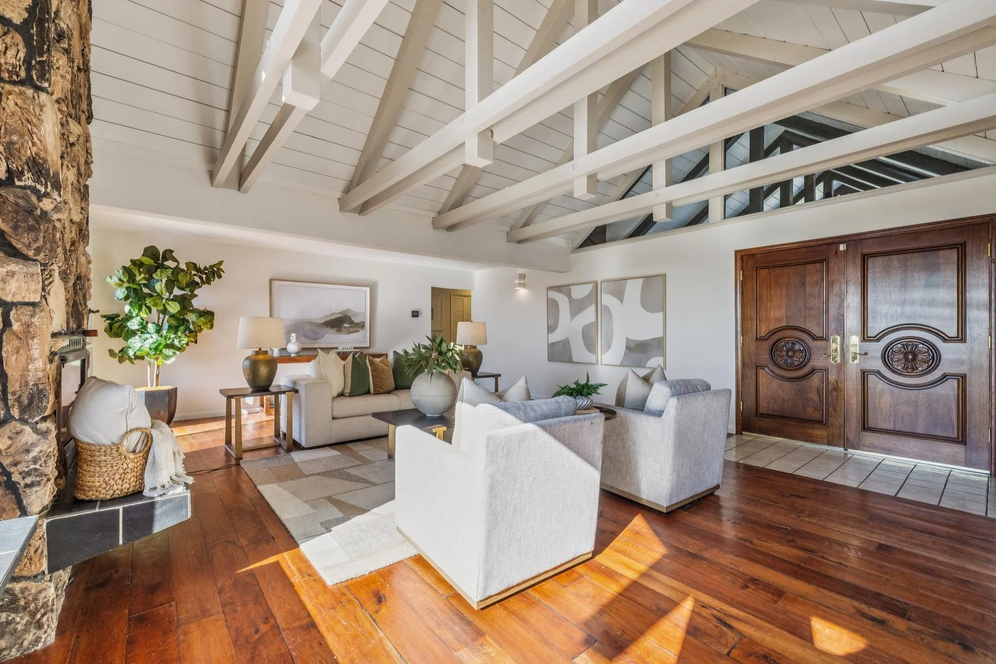 Living room with white sofas, a large leafy plant, framed artwork, wooden double doors, and exposed ceiling beams.