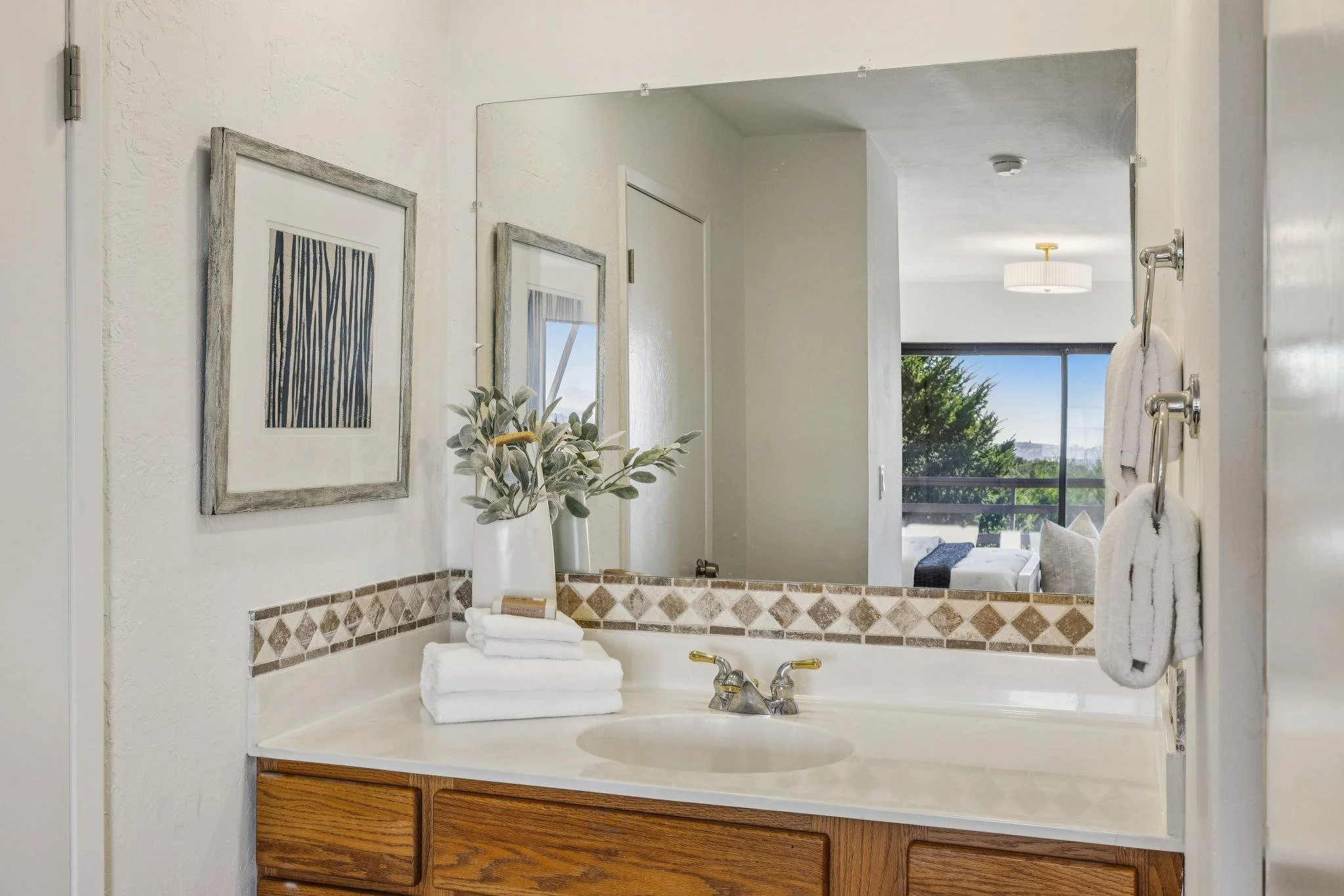 Bathroom sink with a large mirror, framed artwork, a potted plant, folded towels, and a view of a living room with a large window and trees outside.