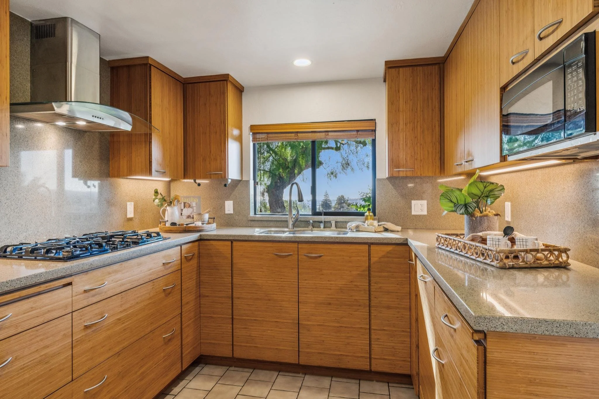 Kitchen with wooden cabinets, granite countertops, a window overlooking trees, a stove with four burners, a backsplash, a plant, and various kitchen items on the counters.