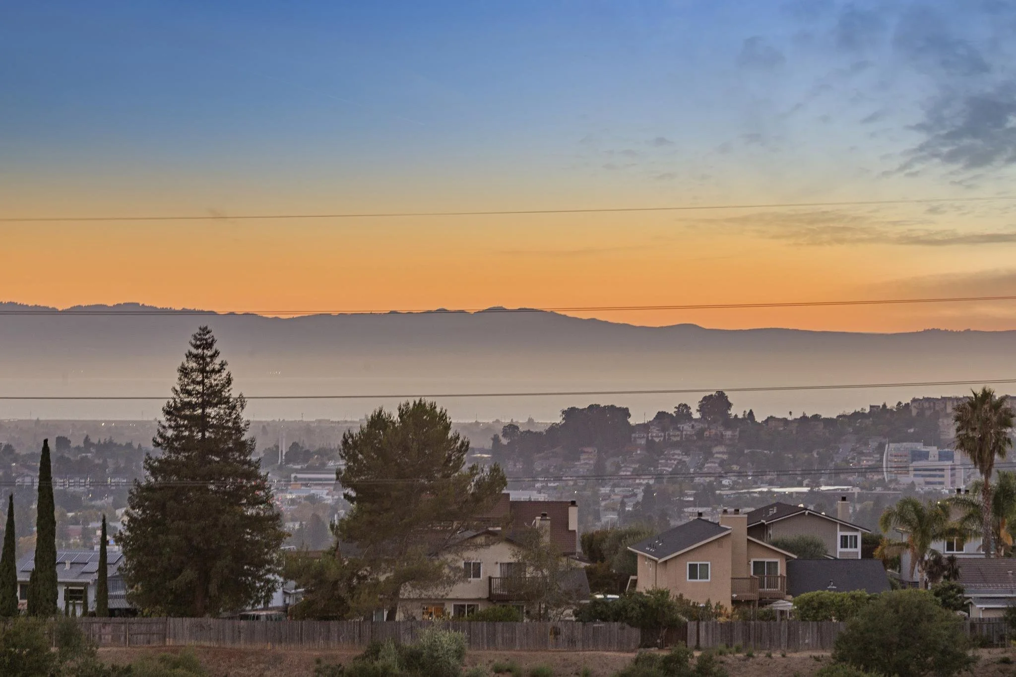 A sunset view over a suburban neighborhood with houses, trees, and a distant mountain range, with power lines crossing the sky.