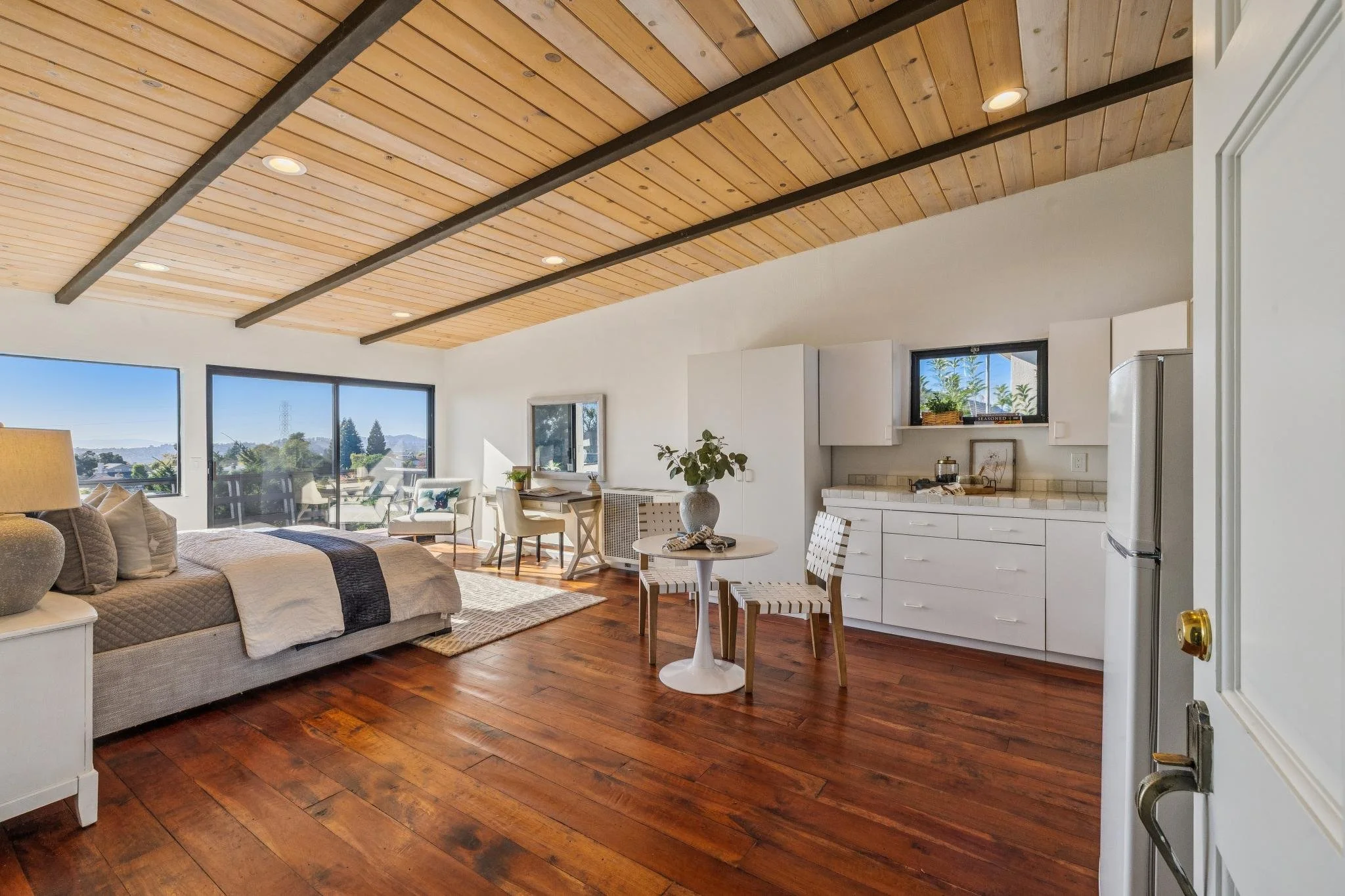Living room with white walls, wooden floor, large windows, a beige couch, a white dining table with chairs, a small white cabinet, and a ceiling with wooden panels and black beams.