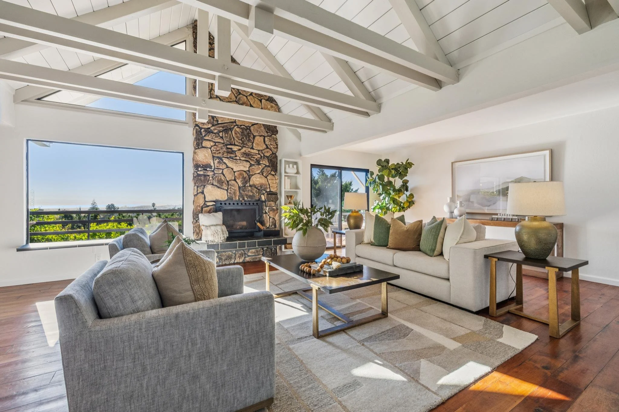 Living room with white ceiling, exposed beams, large windows showing outdoor view, stone fireplace, gray and white couches, wooden and metal coffee table, plants, and lamps.
