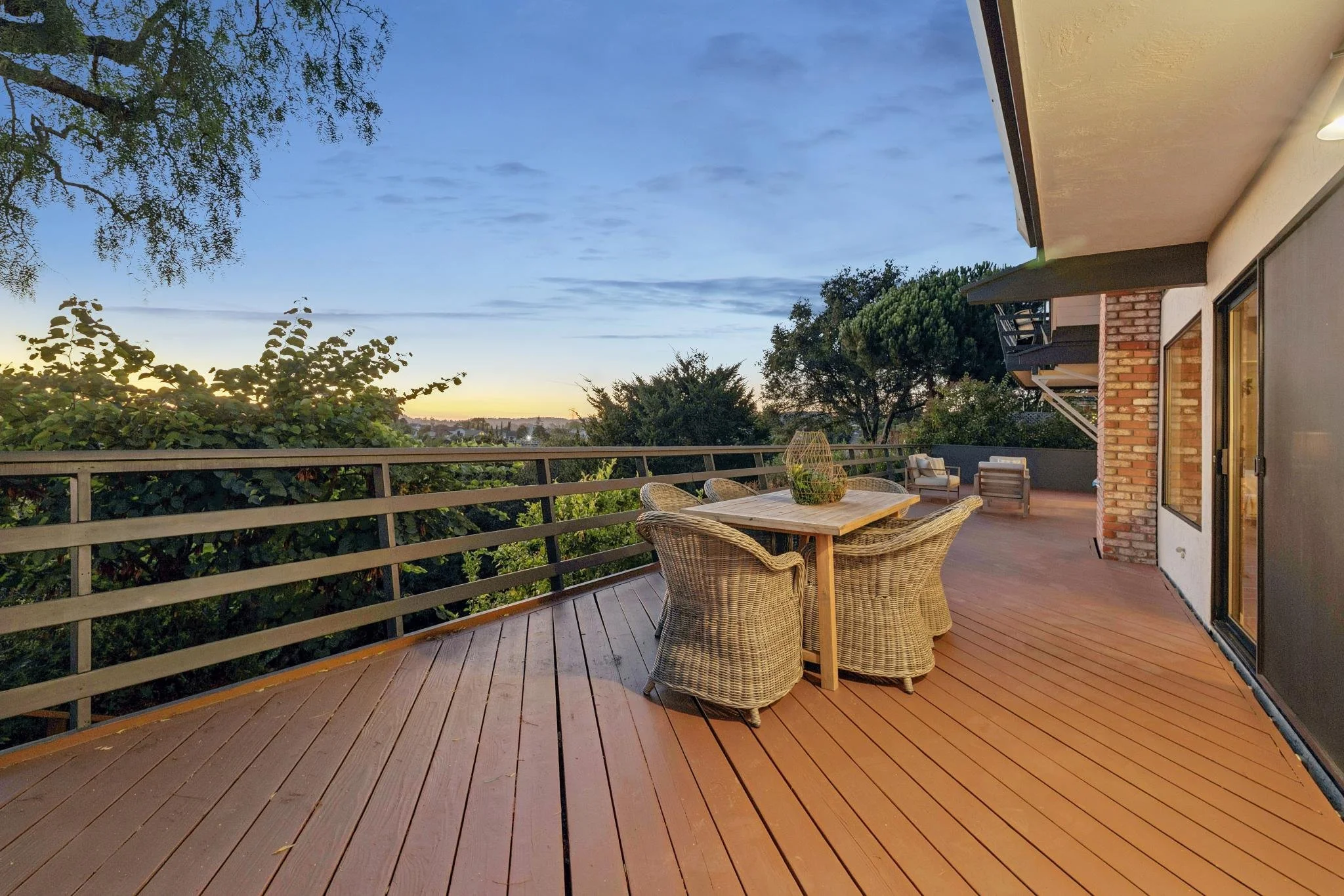 A wooden outdoor terrace at dusk with a round wicker table and chairs, surrounded by a metal railing, overlooking trees and a distant horizon under a partly cloudy sky.