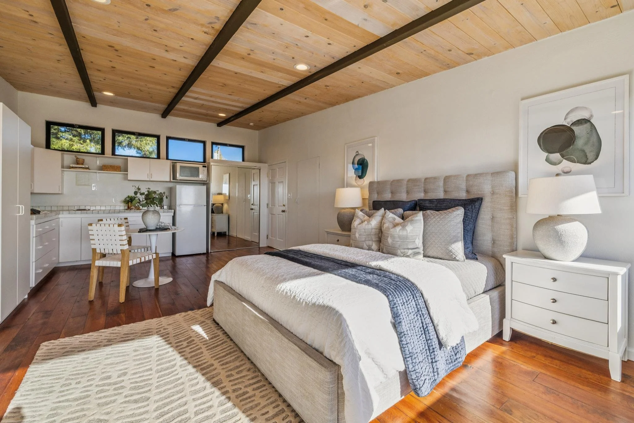 A bedroom with a large bed, white side tables, and a beige upholstered headboard. There are framed abstract artworks on the wall, two bedside lamps, and a dark blanket on the bed. The room has wooden flooring, a paneled wooden ceiling with black beam