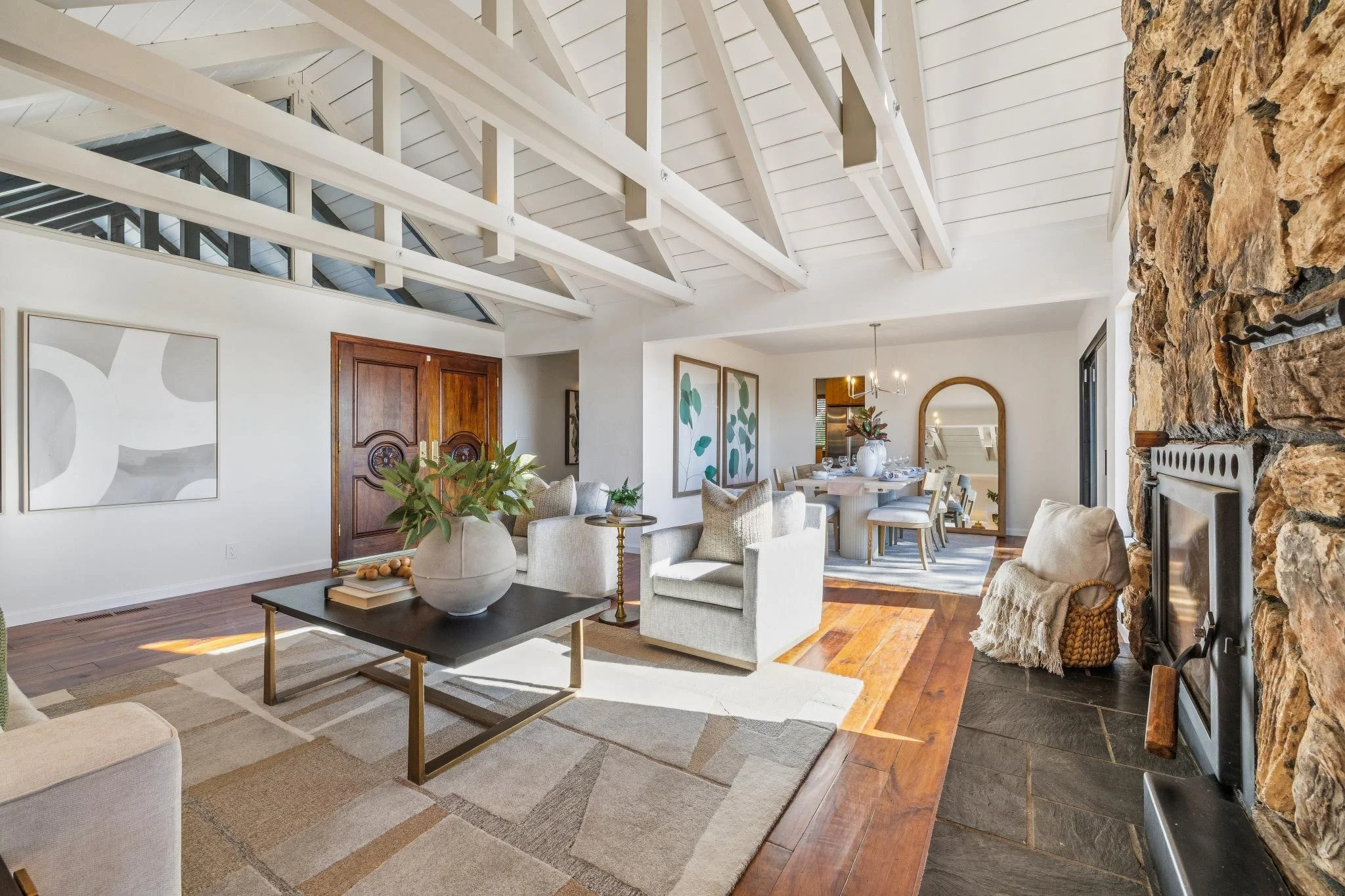 Living room with white vaulted ceiling, exposed wooden beams, stone fireplace, beige armchairs, wooden dining table with chairs, large wall art, and hardwood flooring.