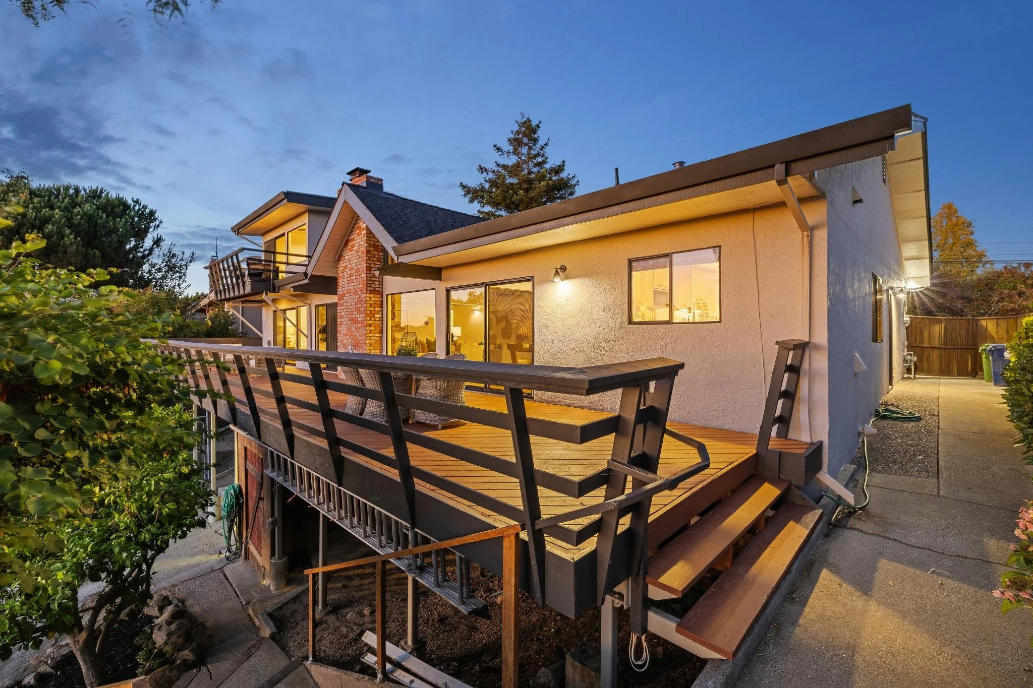 A two-story house with a spacious wooden deck and large windows, illuminated from inside, during dusk.