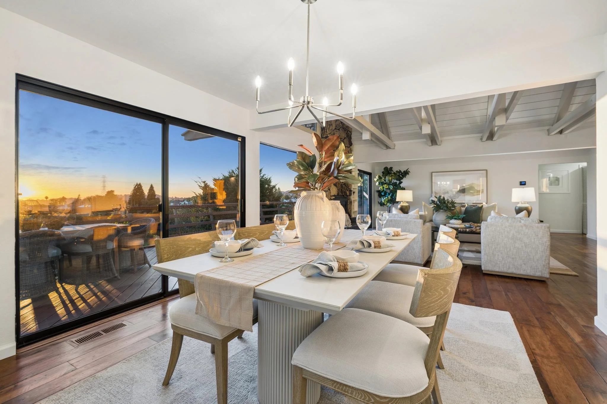 Dining room with a white table set with plates, napkins, and wine glasses, decorated with a large potted plant. Behind the table, there are white upholstered chairs, lamps, framed art on the wall, and a living area with more plants and furniture. Lar