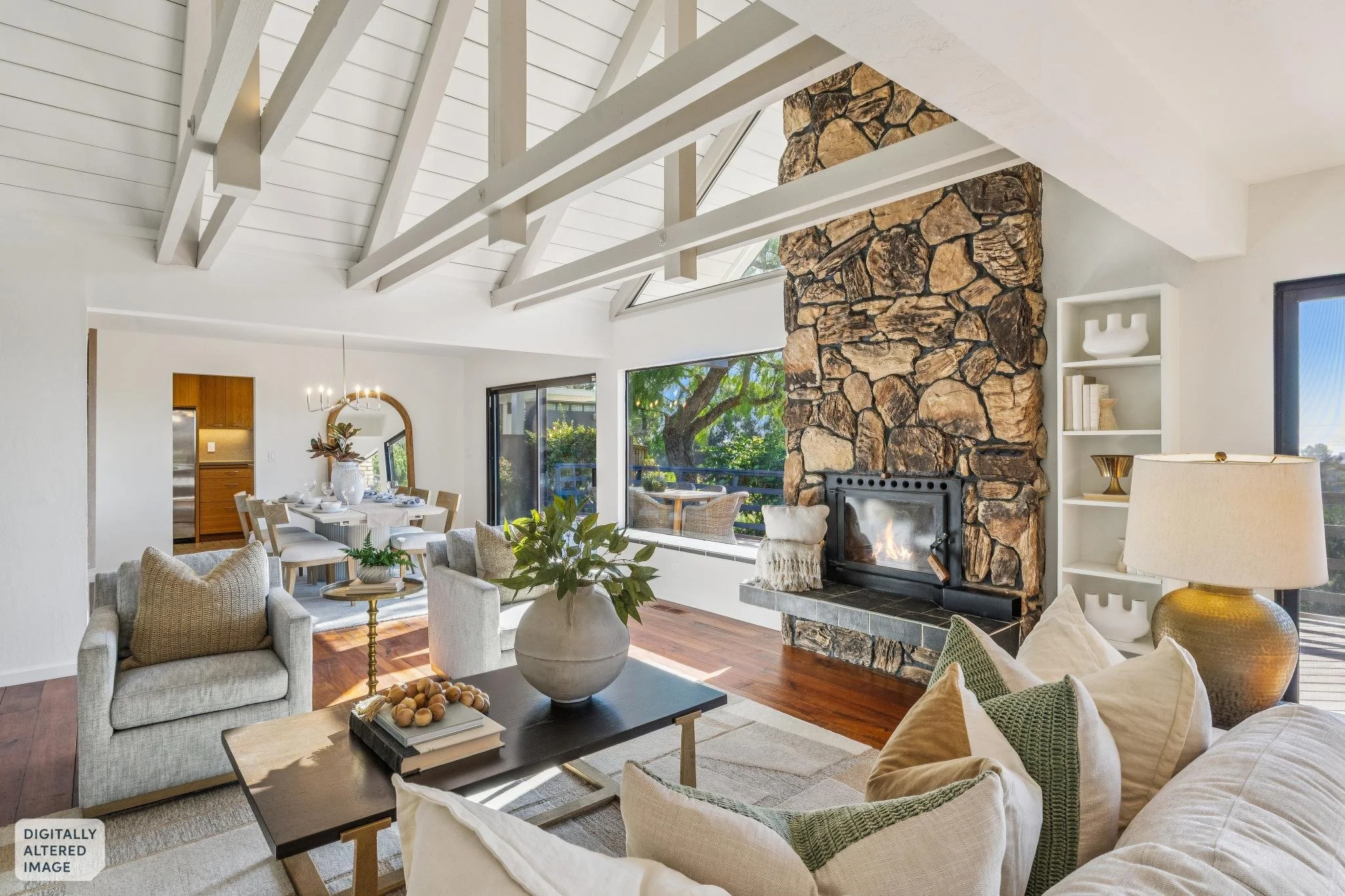 Living room with high, white vaulted ceiling and exposed white beams. A large stone fireplace with a fire is on the right side. Large windows behind the fireplace reveal a tree outside. There is a white bookshelf with decorative items, and a beige la