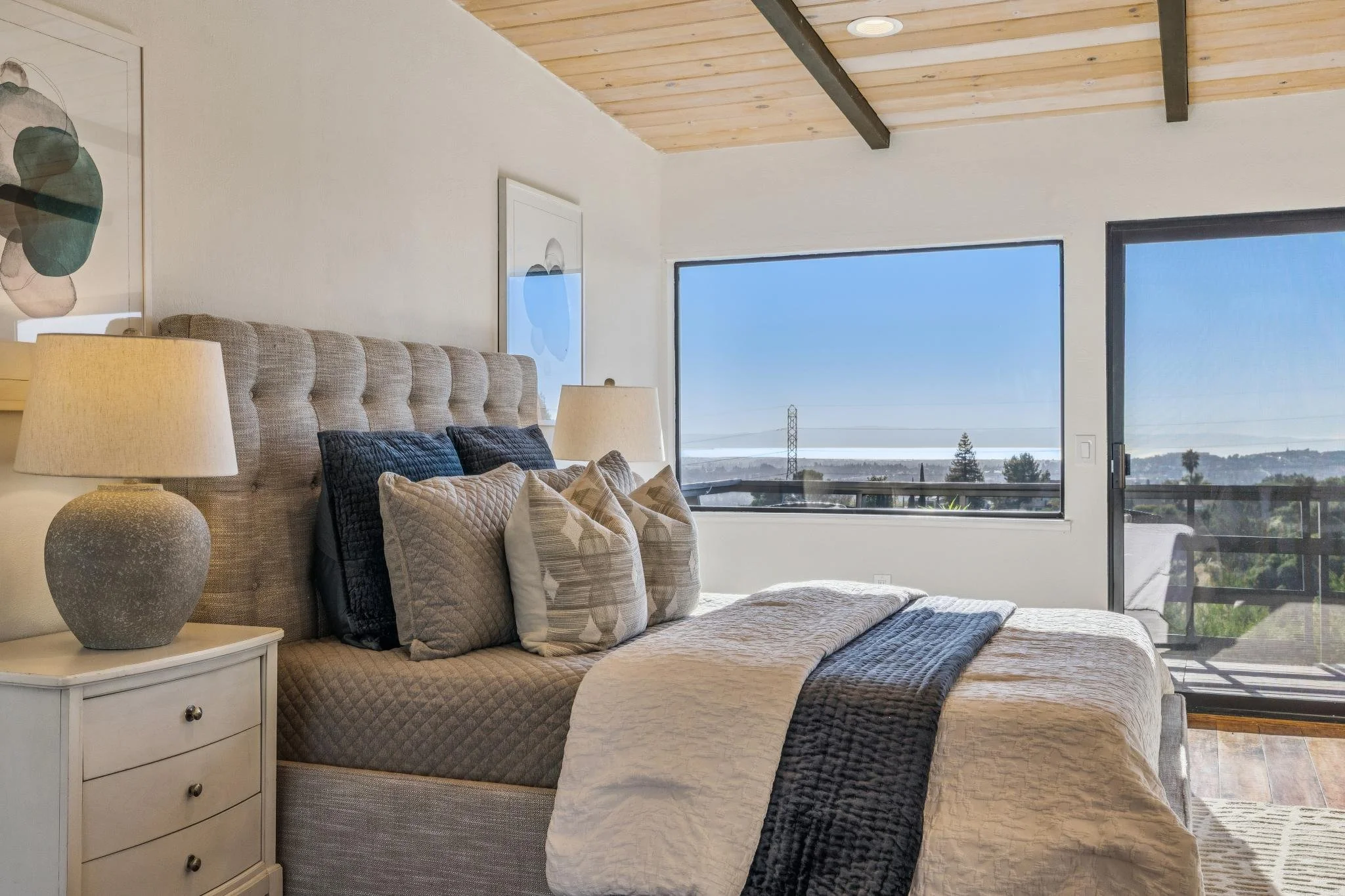 A bedroom with a large bed, multiple pillows, a nightstand with a textured globe-shaped lamp, large windows showing a view of sky and landscape, and a wooden ceiling with exposed beams.