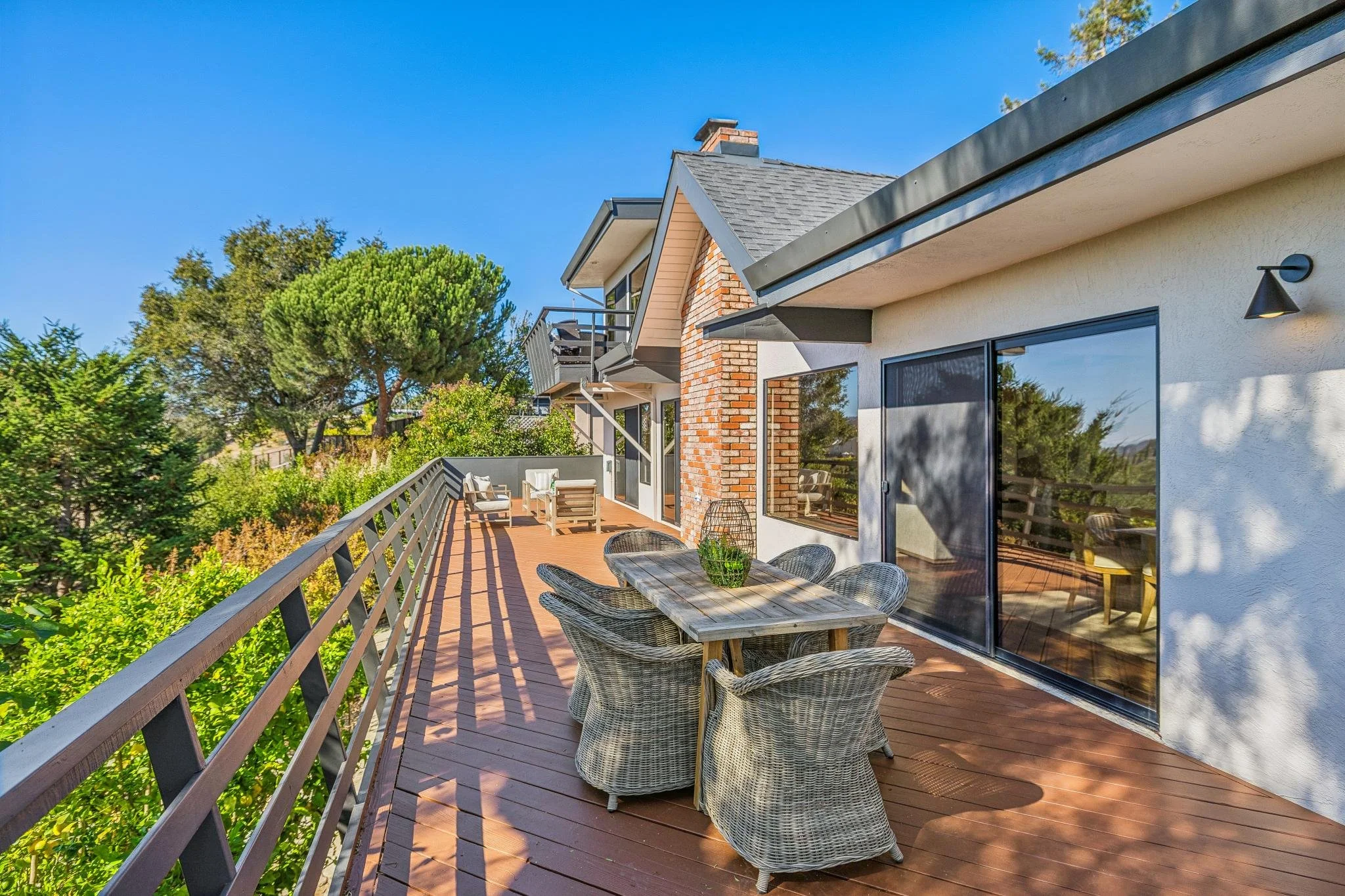 Backyard deck with wicker chairs around a wooden table, large sliding glass doors, exterior wall with brick accents, lush trees and greenery, sunny clear sky