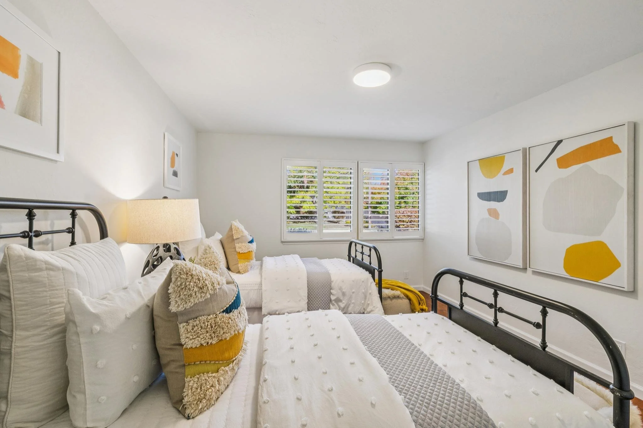 A bedroom with white walls, three black metal-framed beds with white bedding and textured throw pillows, a window with white shutters showing trees outside, and abstract artwork on the walls featuring shapes in yellow, gray, and orange.