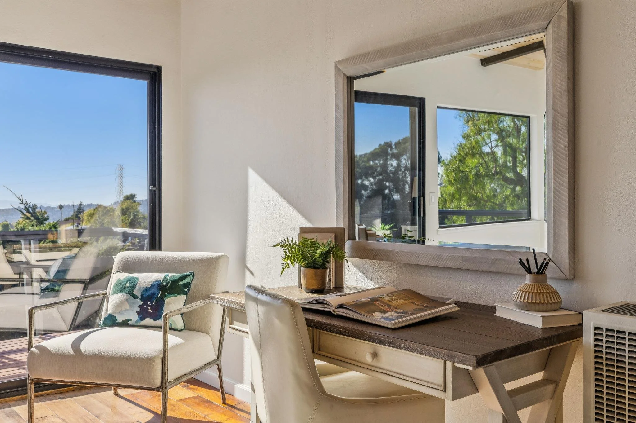A cozy corner of a room with white walls and large windows showing a bright, sunny day with trees and blue sky outside. There is a white wooden desk with open books and a decorative vase with black sticks. Next to the desk is a white upholstered chai