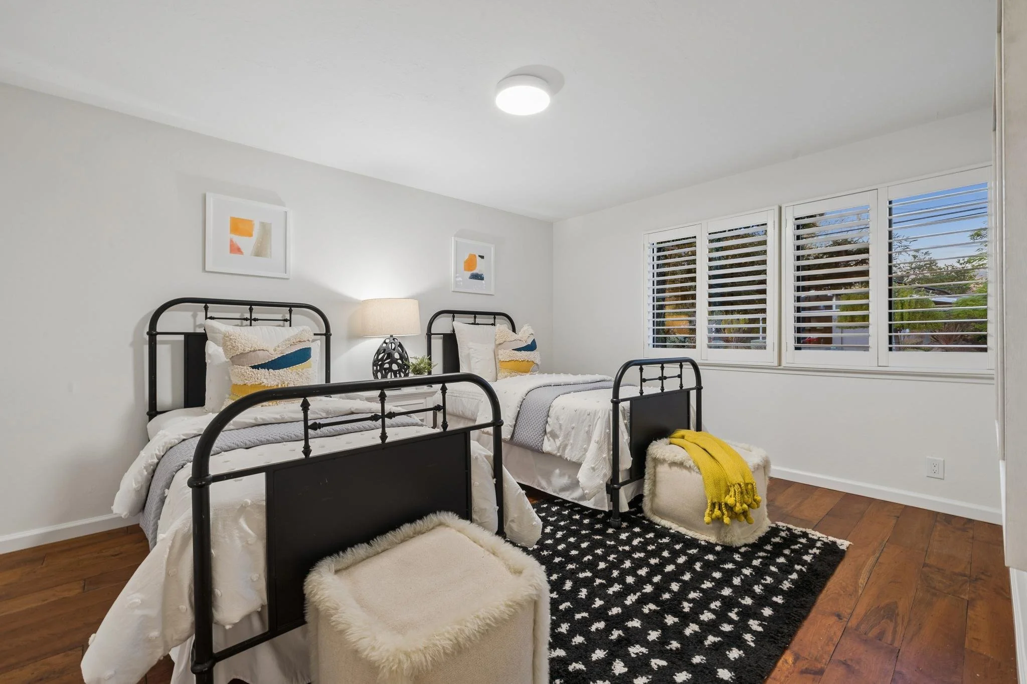 A bedroom with two twin beds, black metal frames, white bedding, colorful pillows, a black and white patterned rug, a beige stuffed ottoman, a fluffy cream chair with a yellow blanket, a window with white blinds, and framed abstract art on the light 