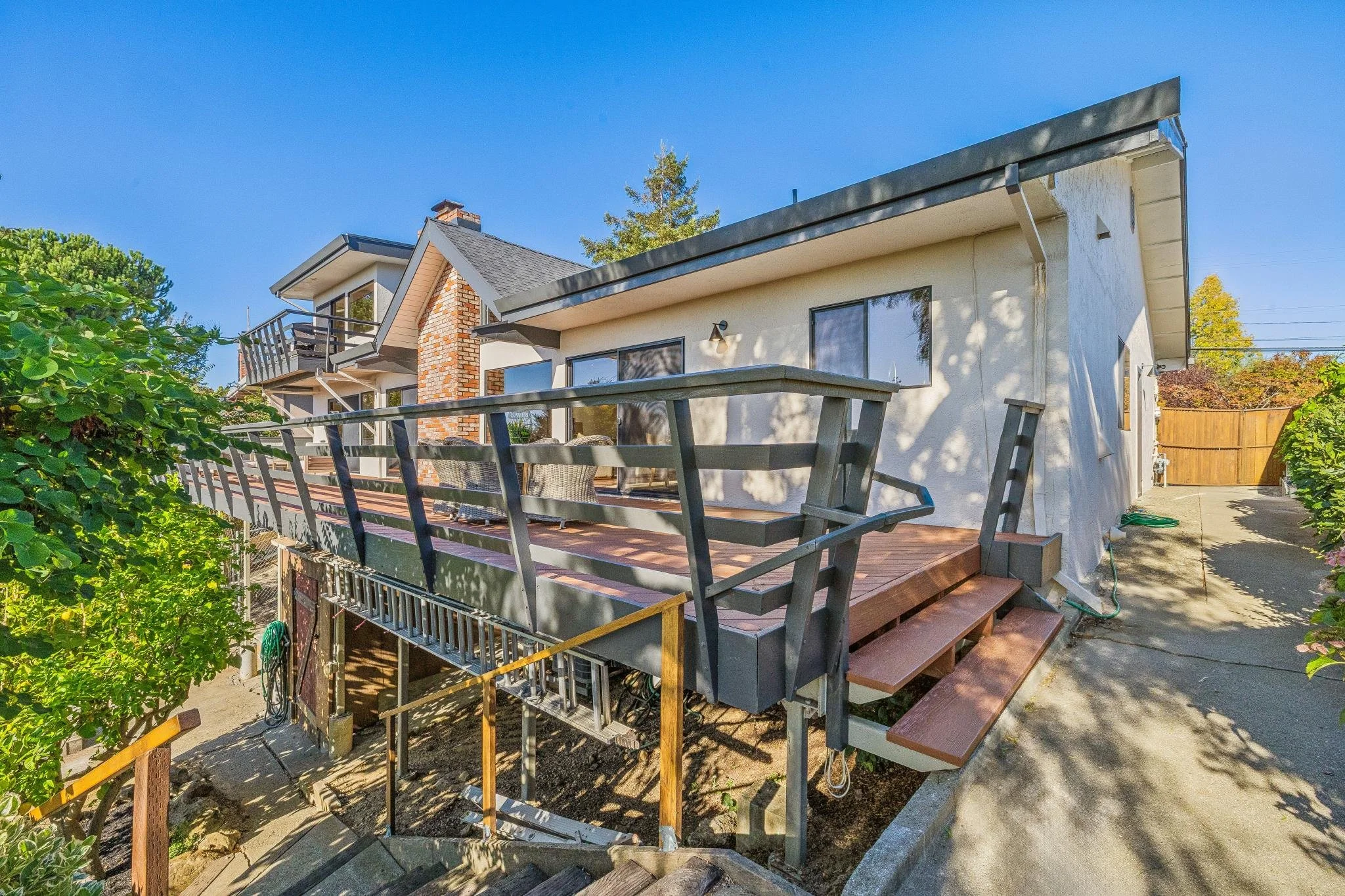 A two-story house with white exterior walls, large windows, and a balcony. There's a wooden deck with steps leading down, and a black metal railing around a portion of the deck. The house has a brick chimney, and there are trees and clear blue sky in