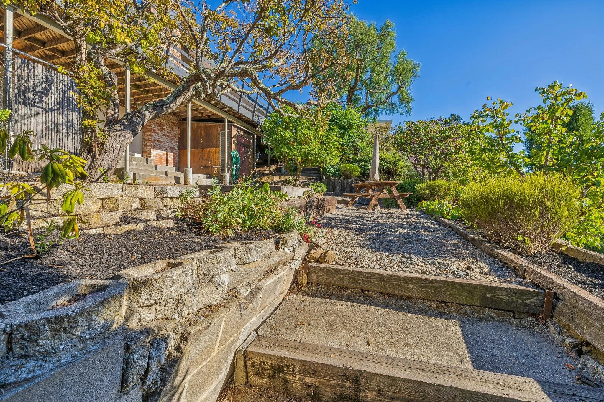 A landscaped backyard with stone stairs leading to a wooden deck, surrounded by green trees, bushes, and an outdoor picnic table with an umbrella on a sunny day.