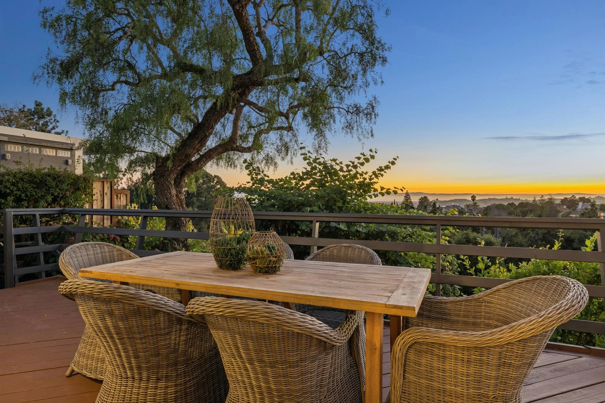A wooden outdoor table with wicker chairs, decorative baskets, and lush greenery on a deck with view of a sunset over a distant landscape of trees and water.