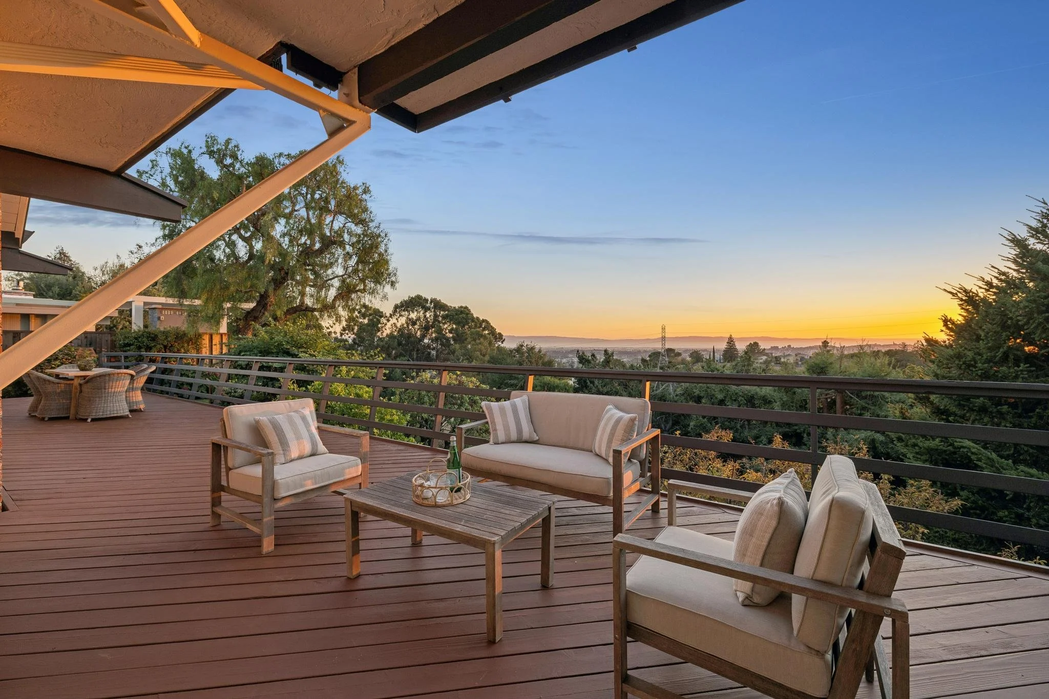 A spacious outdoor balcony at sunset with comfortable patio furniture including cushioned chairs and a coffee table, overlooking trees and a distant horizon.