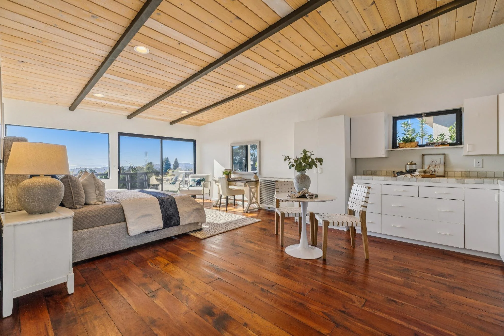 Living room with a vaulted wooden ceiling with black beams, large windows showing an outdoor view, and hardwood floors. Furniture includes a light-colored sofa, white side table with a lamp, a small round dining table with chairs, and a white kitchen