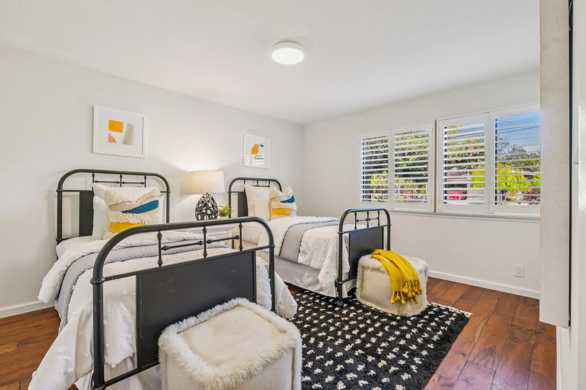 A bedroom with two beds featuring black metal frames, white bedding, and decorative pillows. A fluffy white footstool with a yellow blanket is beside one bed. There are framed abstract art pieces on the white walls, a window with white shutters letti