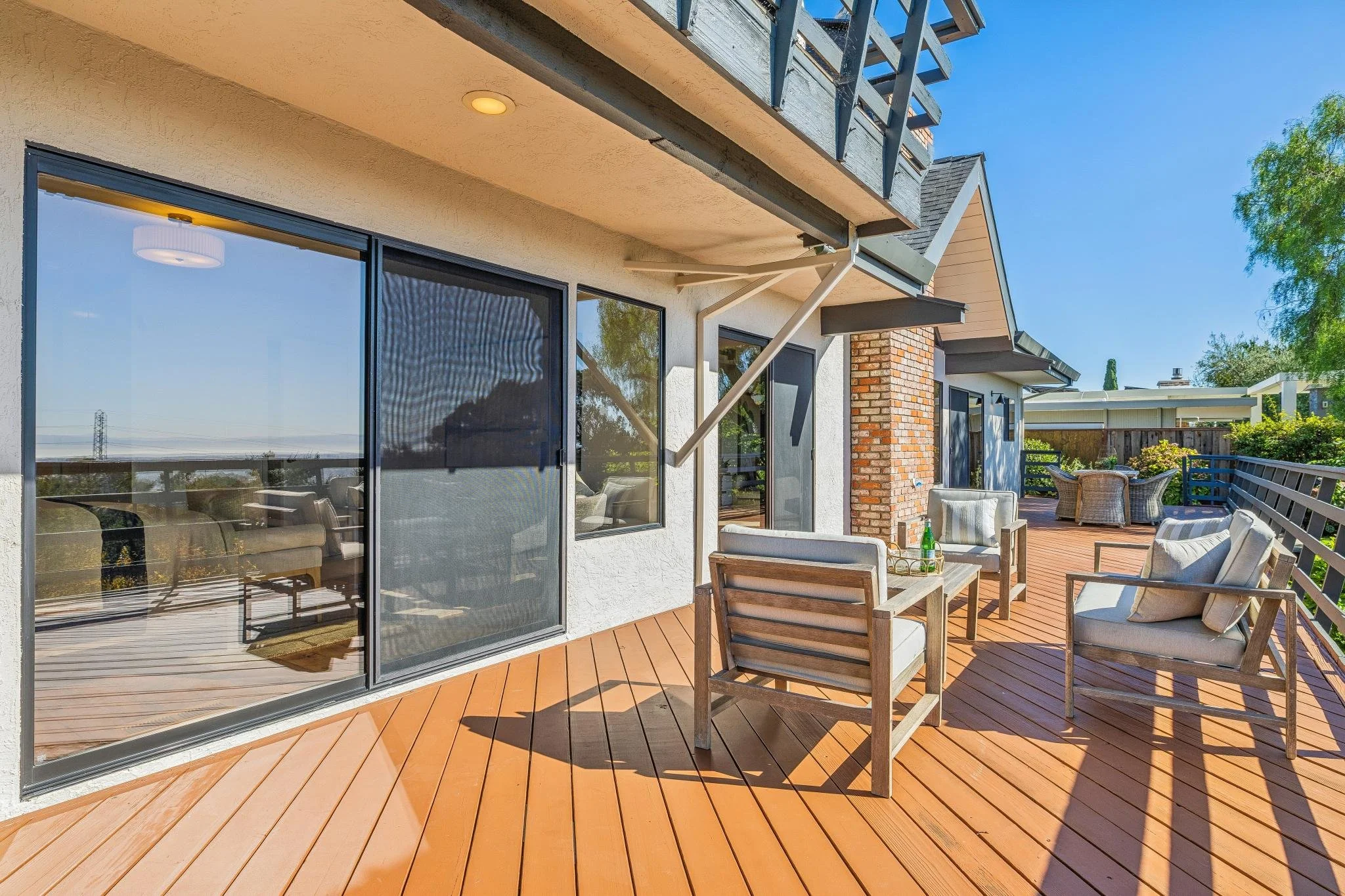 Backyard patio with wooden decking, outdoor furniture, and large sliding glass doors reflecting the blue sky and nearby trees.