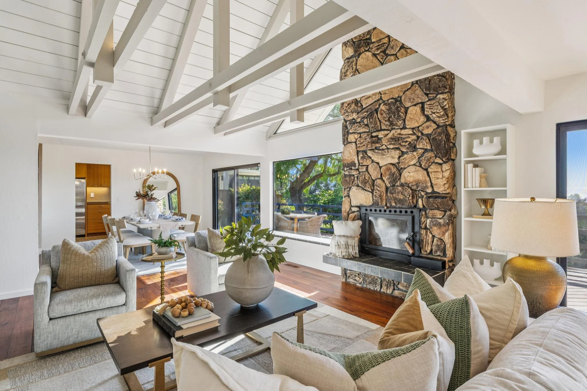 Living room with a stone fireplace, white ceiling beams, large windows, and a dining area in the background. Furniture includes armchairs, a sofa, a coffee table with decorative items, and a large vase with plants. Neutral tones and natural light cre