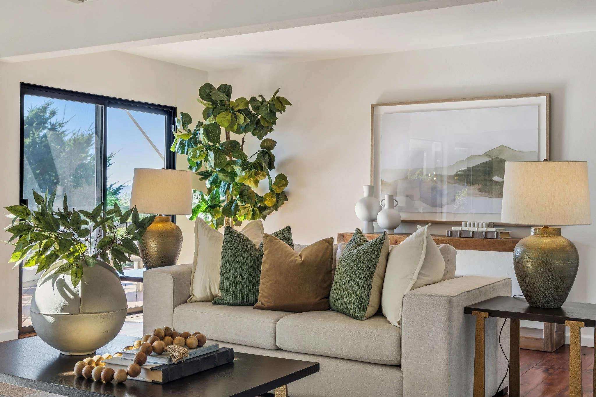 Living room with large potted plants, a white sofa with multiple pillows, and a coffee table with decorative items, near a window showing trees outside.