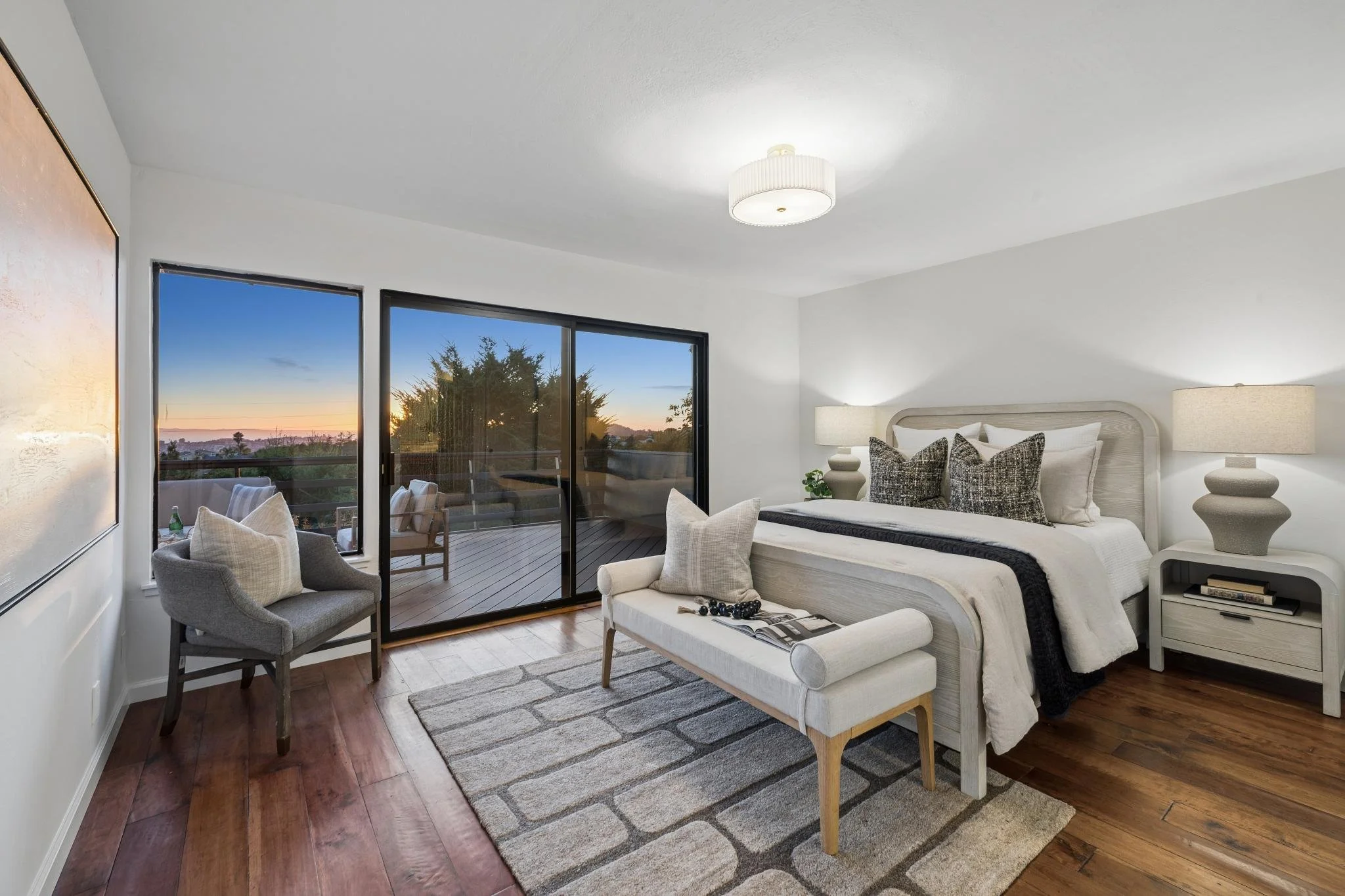 Bright bedroom with a large bed, nightstands with lamps, and a sliding glass door leading to a balcony showing a sunset view with trees.