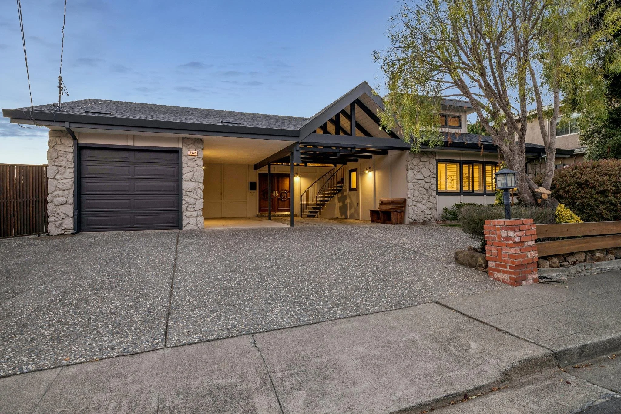 Modern house with stone and stucco exterior, garage door, front porch with stairs, garden, trees, and a lamp post, taken during dusk or evening.