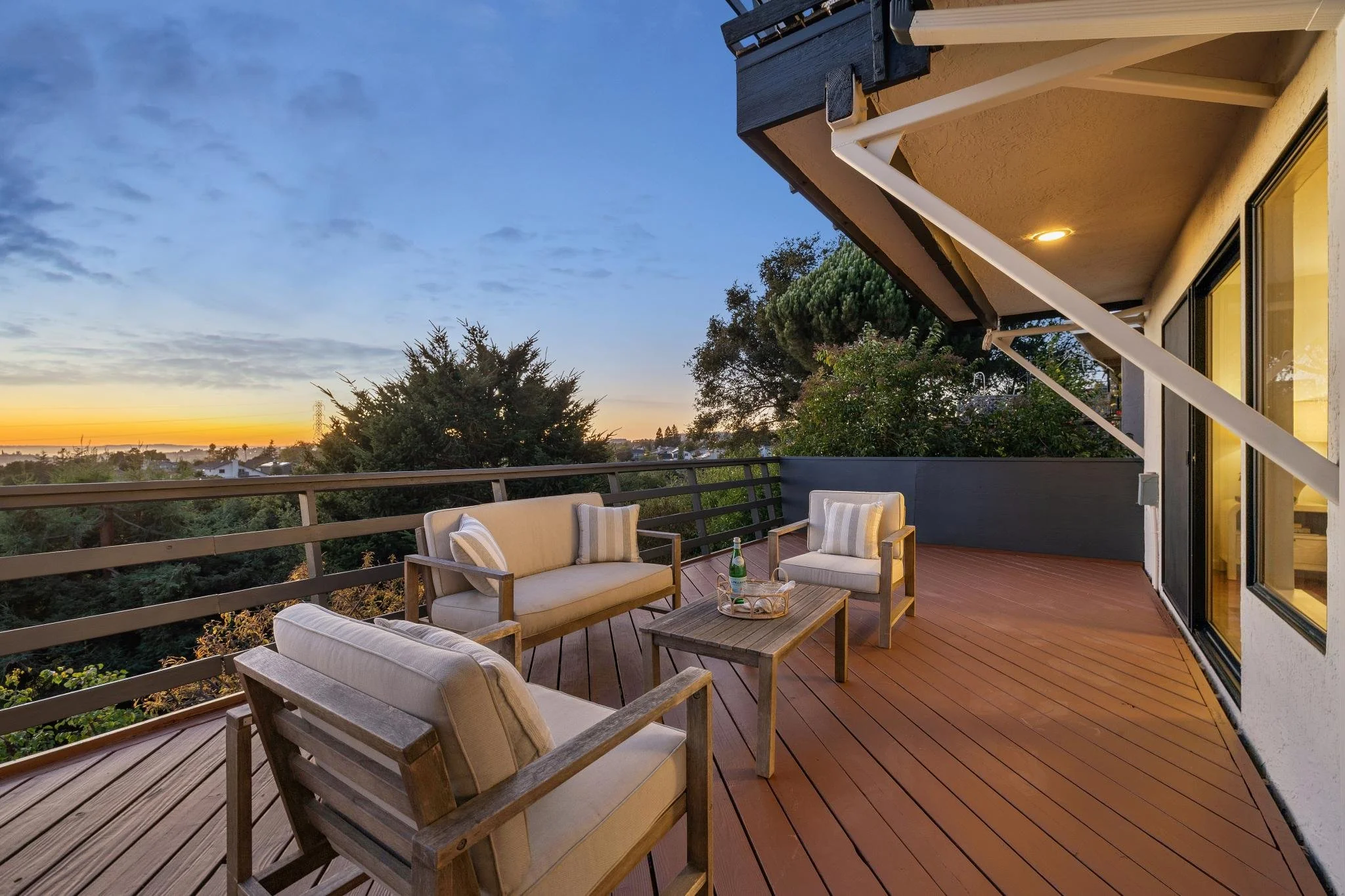 An outdoor balcony at sunset with seating and a view of trees and the horizon.