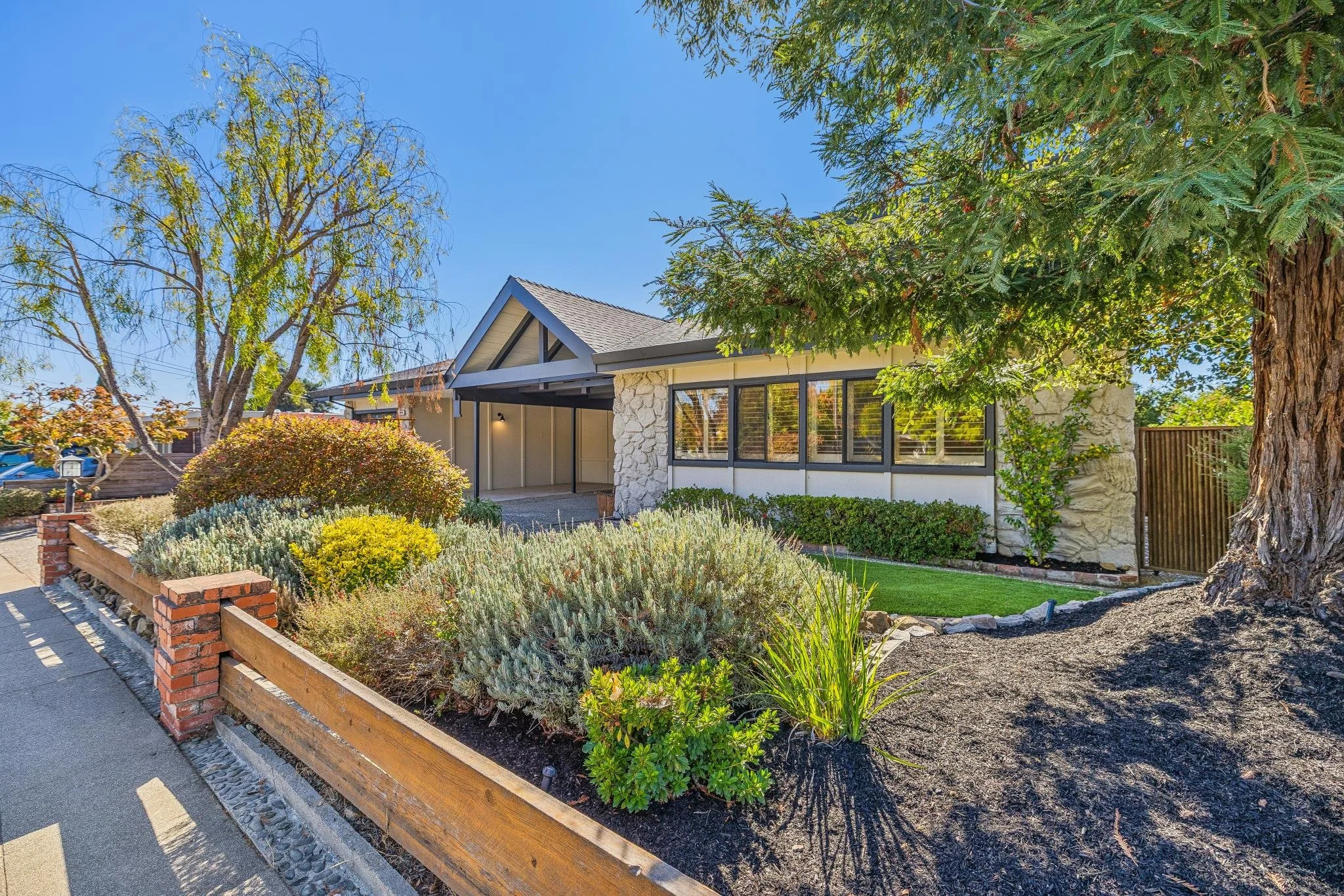 A suburban house with a front yard featuring a variety of shrubs, bushes, and trees, with a sidewalk and a low brick and wood fence in the foreground. The house has a modern design with large windows and white stone accents, under a clear blue sky.